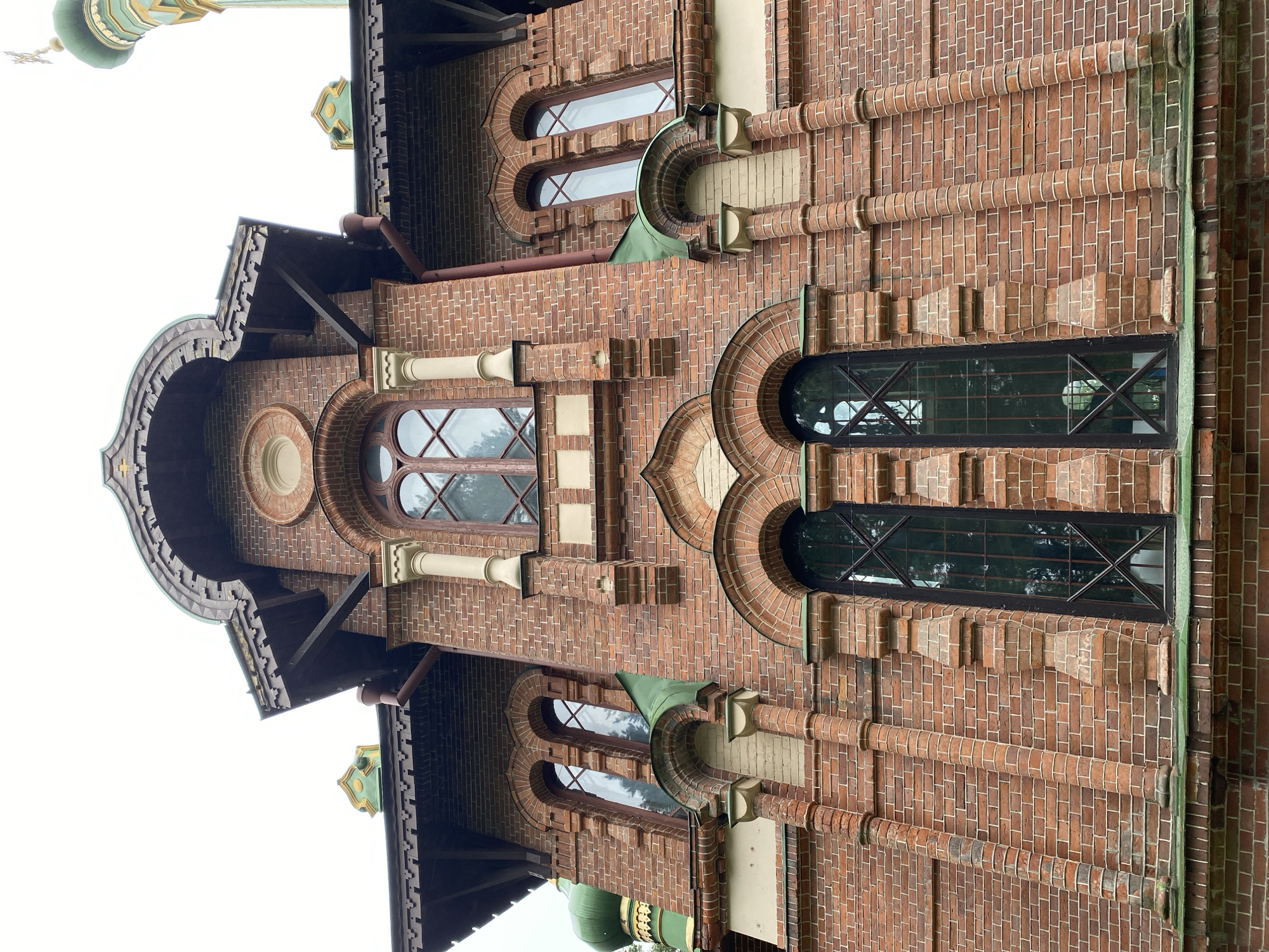 The image shows the facade of a brick building, possibly a church or historical structure, located in Priozersk, Russia. The building features red brick construction with decorative architectural elements. Prominently visible are arched windows with dark frames and intricate metal grates. The roofline is complex, with wooden eaves adorned with decorative carvings. Green domes, typical of Russian Orthodox churches, are visible on the top of the structure, one with a cross. The sky is overcast. The perspective is from a low angle, looking up at the building.