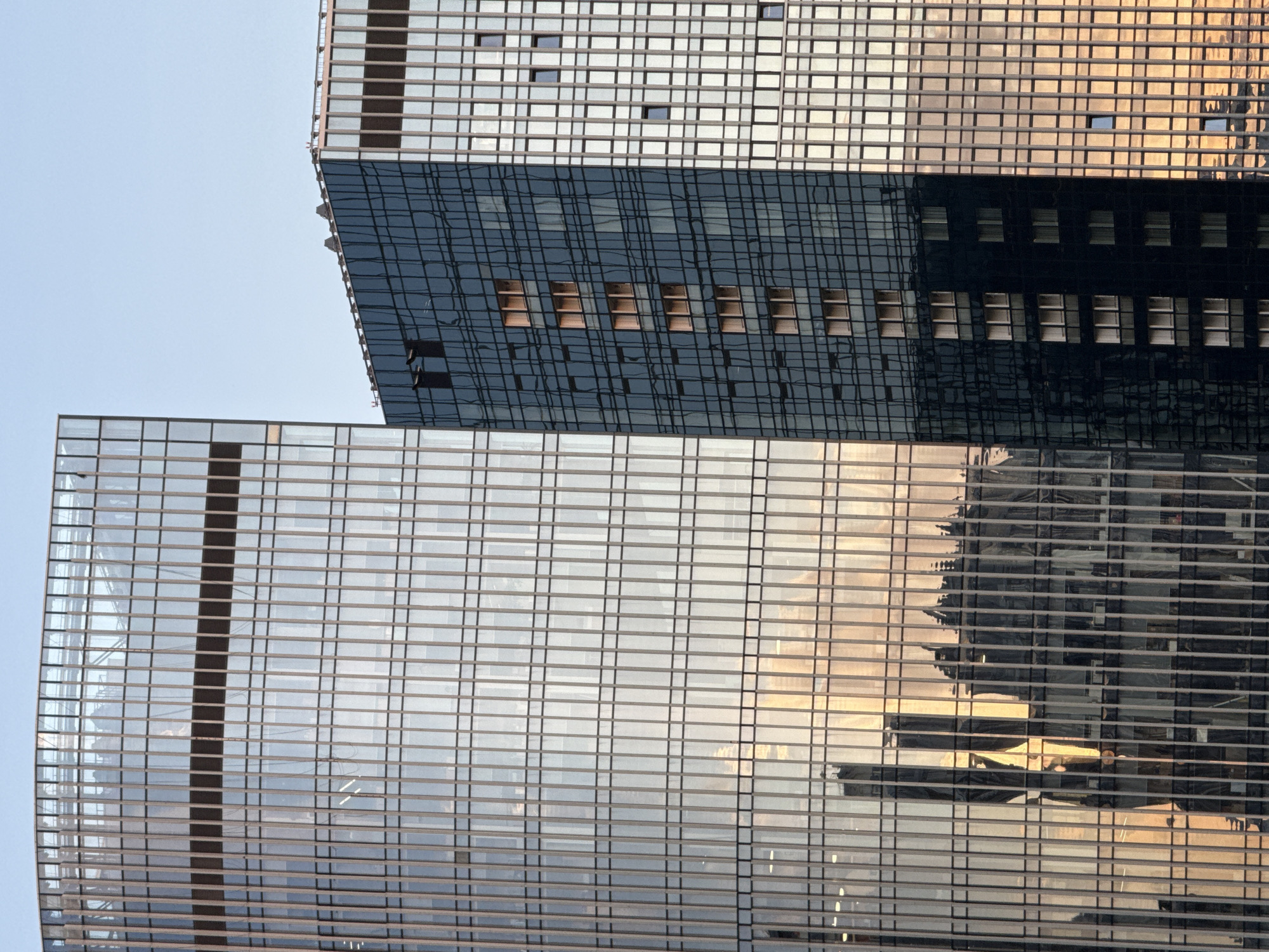 This media file shows a close-up, vertical-orientation view of modern glass skyscrapers in Khimki, Russia. The composition is dominated by the geometric patterns of the buildings' facades. The upper portion of the image features a section of a building with a lighter-colored exterior, possibly made of metal panels and glass, showing a grid of small, dark square windows. Below this, a darker section of a skyscraper with a reflective, dark glass facade is visible. This dark glass reflects the warm, golden light of the sky, creating an impression of sunset or sunrise. The reflection reveals the grid pattern of windows on another building, some appearing as lighter rectangles within the darker glass. The lower portion of the image is filled with a large section of a building clad in light-colored, highly reflective glass. This glass also reflects the sky and surrounding structures, with a vertical dark band running down its center, breaking the horizontal lines of the windows and panels. The reflections in this lower section are more diffused and show subtle shifts in color, suggesting the presence of clouds or the changing light. The overall scene evokes an urban environment with a focus on architectural design and the interplay of light and reflection on glass surfaces. There is no visible text in the image. The time of day appears to be either early morning or late afternoon, given the warm lighting. The weather seems clear, indicated by the bright sky.