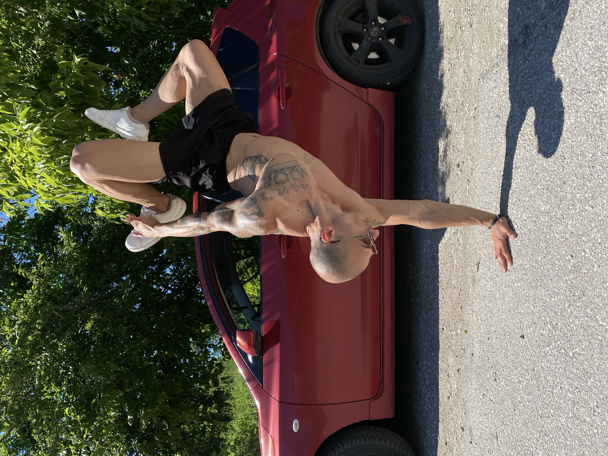 A bald man with tattoos is posing on a red car outdoors. He is wearing black shorts and white sneakers. His legs are inside the car, while his upper body is extended outwards with his arms outstretched. The car is parked on a paved surface, with trees and vegetation visible in the background. The location is likely in Pleven, Bulgaria.