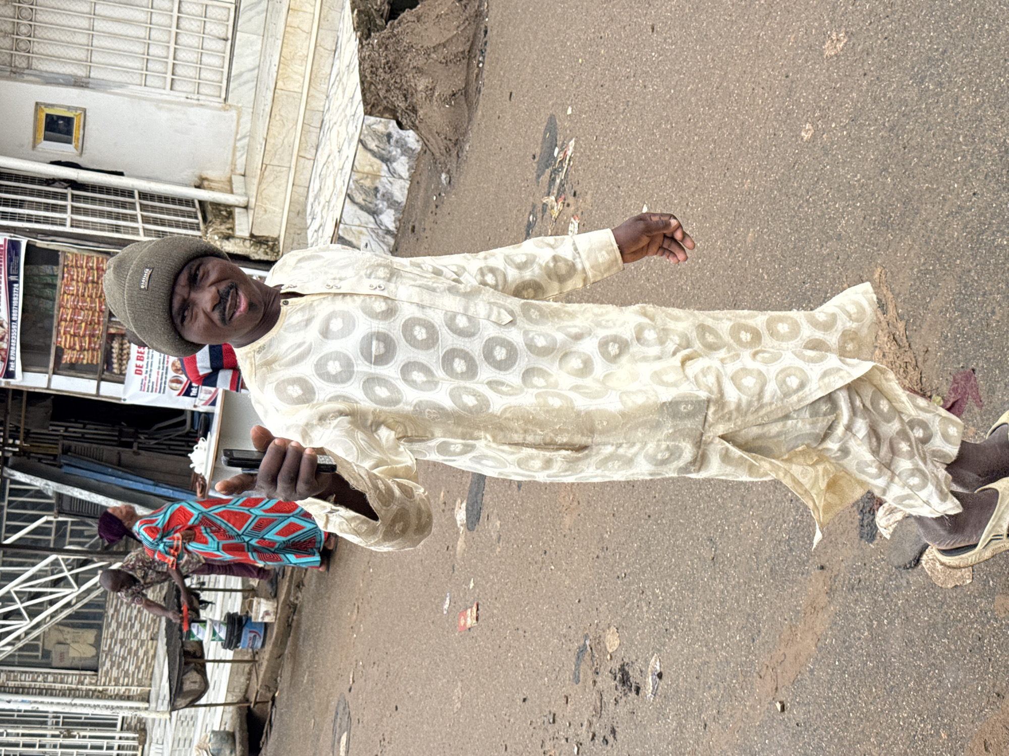A man wearing a knit cap and a light-colored patterned robe stands on a paved street. He is looking towards the camera with a slight smile and holding a dark-colored object, possibly a phone, in his left hand. In the background, to the left, a building with white grating and a storefront is visible. Another person wearing a colorful patterned dress can be seen in the background, walking away from the camera. The ground is asphalt with some scattered debris. The scene appears to be outdoors in what seems like an urban or market setting. The lighting suggests it is daytime. The visible text on a sign in the background reads "DE BE".