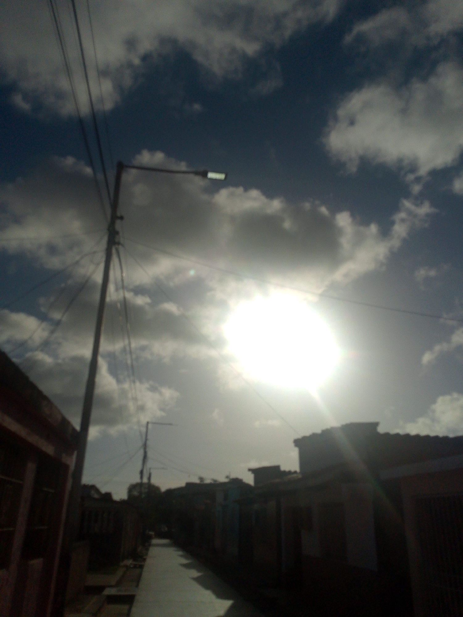 In the city of Juan Griego, Venezuela, a street scene unfolds under a bright, cloudy sky. The sun is a powerful, luminous presence in the upper right quadrant of the image, casting a strong glare and creating lens flare effects. A tall utility pole with numerous wires stretches vertically from the lower left to the upper center, its silhouette sharp against the sky. To the left, a portion of a building with a reddish-brown roof and dark walls is visible. A paved pathway, appearing to be a street or sidewalk, leads into the distance, bordered by more buildings on either side, which are mostly in shadow and difficult to discern in detail. The architecture suggests a residential or commercial area. The overall impression is one of a bright, possibly hot, day in a populated urban environment.