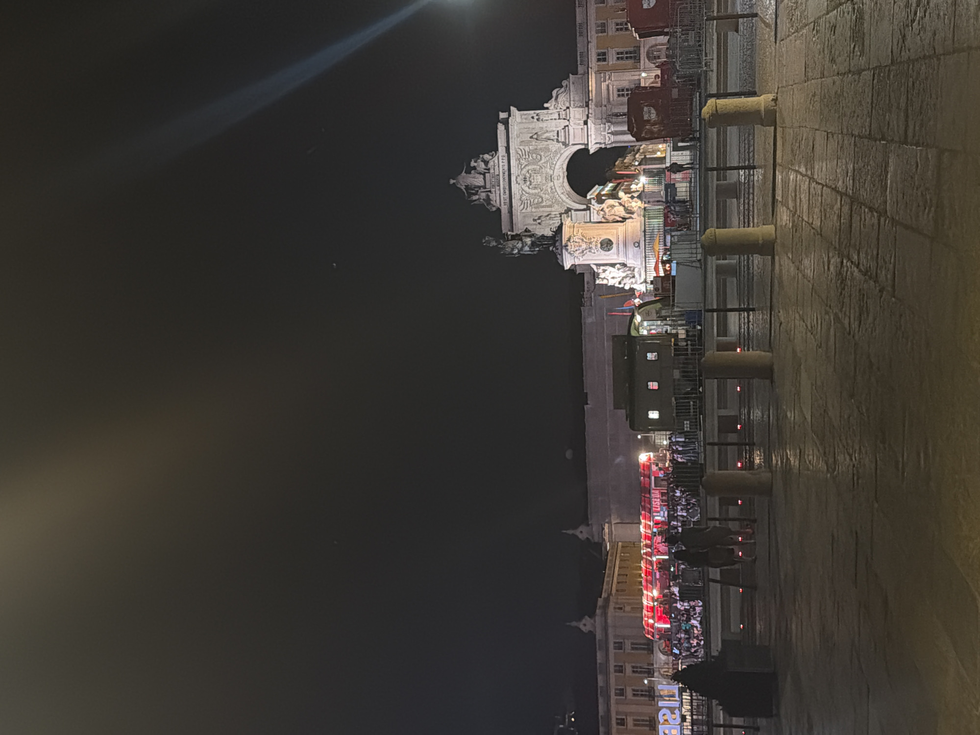 The image captures a night-time view of Praça do Comércio in Lisbon, Portugal. Dominating the background is the Arco da Rua Augusta, a triumphal arch illuminated by artificial lights. In front of the arch, a statue of King José I on horseback stands on a pedestal. The square itself is paved with stone, and the ground appears wet, reflecting the ambient light, suggesting recent rain.
In the mid-ground, a bright red double-decker bus, labeled "LISBOA," is visible, surrounded by a crowd of people. Various temporary structures and stalls are set up in the area, some adorned with lights and banners. Several people are visible in the foreground and mid-ground, including a couple walking away from the camera in the left foreground. Numerous bollards line the edge of the pedestrian area. The sky is dark, with a faint light beam visible in the upper right corner.