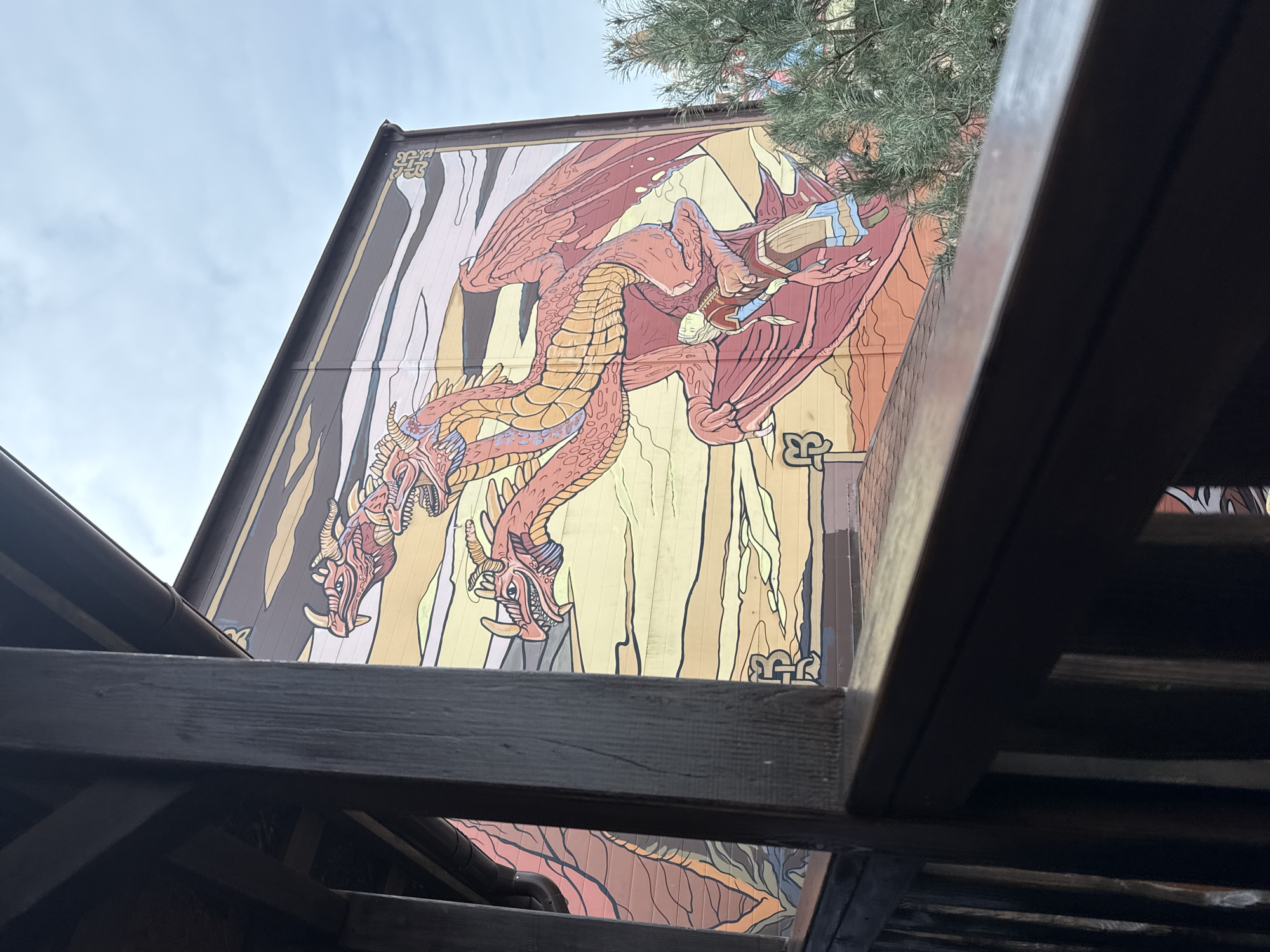 A large, colorful mural featuring a fantasy theme is prominently displayed on the exterior wall of a building within Sochi Park, Sochi, Russia. The central element of the mural is a three-headed dragon, rendered with red-orange scales, large wings, and horned heads with visible teeth. This dragon is depicted holding a human figure, dressed in a beige tunic with blue accents, in its left claw. The mural's background consists of stylized horizontal bands of yellow, orange, and brown, with intricate Celtic knot patterns integrated into the design borders. A dark brown gutter runs along the top edge of the building wall. The scene is viewed from an elevated perspective, with dark, weathered wooden posts and a railing of a nearby structure occupying the immediate foreground and the left side of the frame. Above the building, a light grey, cloudy sky is visible. A portion of a green coniferous tree is present on the far right, partially obscuring the lower right section of the mural.
