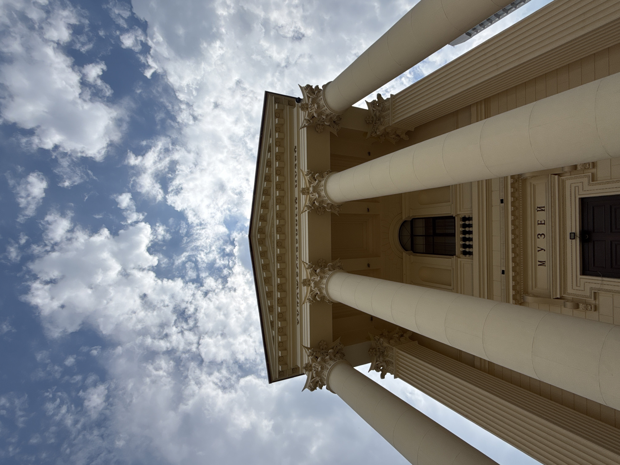 A low-angle shot captures the imposing facade of the Museum of the Local History in Sochi, Russia, against a backdrop of a dynamic sky filled with white, puffy clouds. The building is a grand, neoclassical structure characterized by its large, cream-colored Corinthian columns that dominate the foreground. These columns, with their fluted shafts and ornate capitals, are strategically placed to create a sense of depth and scale, leading the eye upward towards the building's pediment.
Above the columns, a decorative entablature is visible, featuring intricate carvings and moldings. A prominent window with a dark frame and a decorative railing is set into the upper facade, hinting at the interior. Below this, the word "МУЗЕЙ" (Museum in Russian) is inscribed in large, dark letters above a large, dark wooden doorway. The overall impression is one of historical significance and architectural grandeur. The lighting suggests it is daytime, with the sun casting subtle shadows on the building's features. No people are visible in the frame. The perspective emphasizes the verticality and scale of the museum.