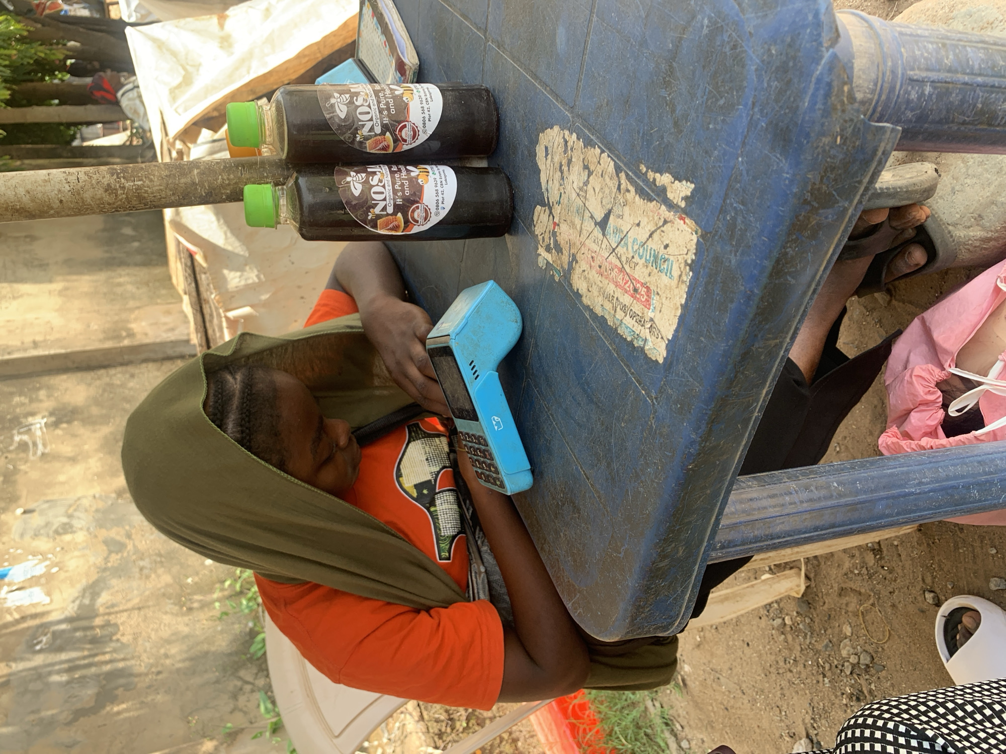A young Black female, wearing an orange t-shirt and a green headscarf, is seated at a blue plastic table. Her head is bowed as she operates a blue card reader. On the table, to her right, are two dark bottles with green caps, each bearing a label with the word "NOSJA" and images of bees. A book or ledger is also on the table. The table is weathered and appears to be outdoors, with a sandy ground and some vegetation visible. In the background, there are structures with corrugated roofing and a woman in white clothing with a red headscarf can be seen further in the distance. The scene appears to be an informal market or roadside stall, possibly in the city of Abuja, Nigeria, given the context.