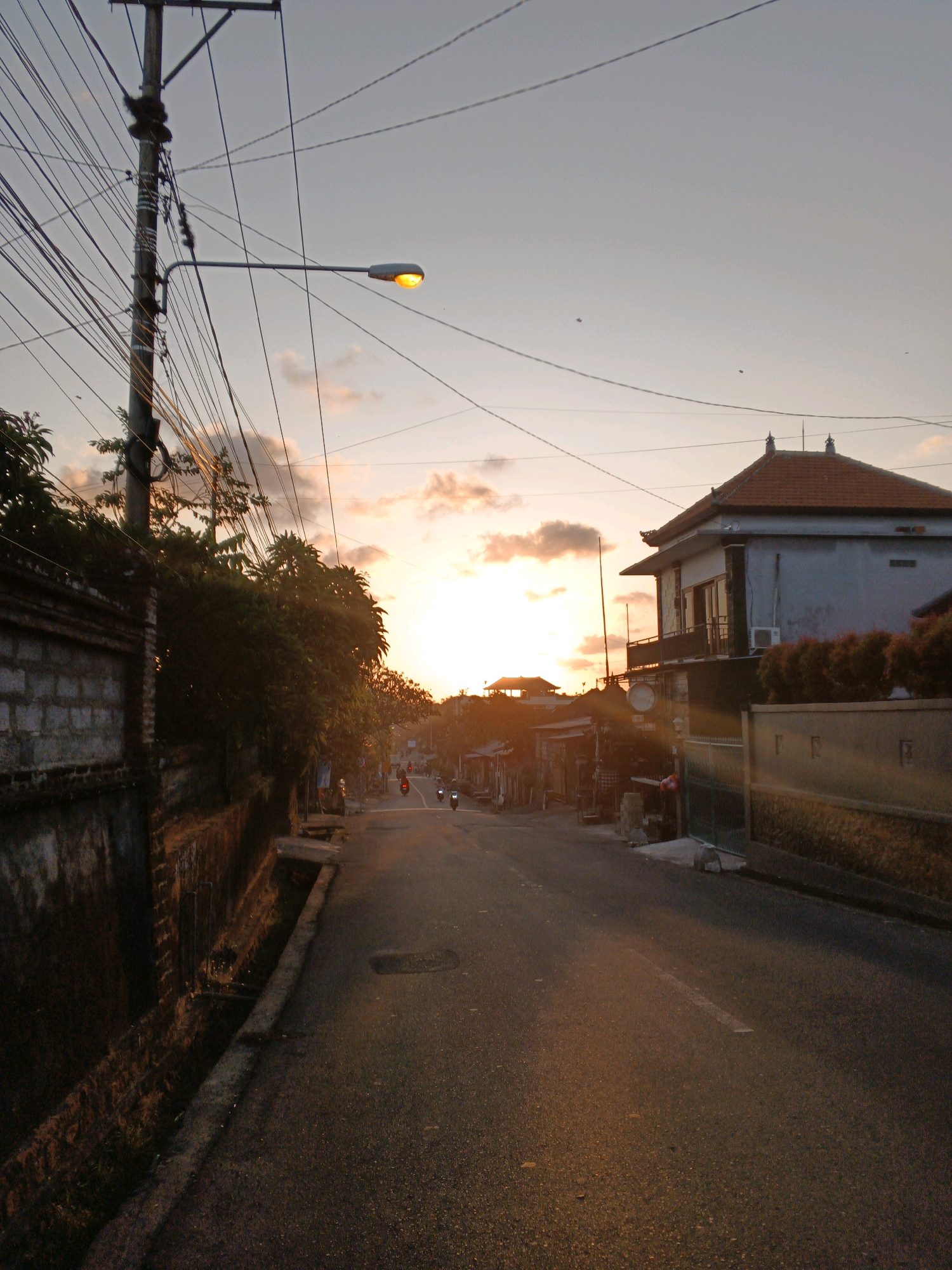 The image depicts a street scene at dusk in Denpasar, Indonesia. A utility pole with numerous electrical wires dominates the left side of the frame, with a street lamp emitting a warm, orange light. To the right, a two-story building with a terracotta roof stands prominently, its facade painted a light blue-gray. The street itself is asphalt, with a faded white line marking the center. The warm glow of the setting sun, visible through scattered clouds, casts a golden hue on the sky and illuminates the buildings and road. On the street, a few motorcycles are visible, one with its brake lights illuminated red. The left side of the street features a low, weathered brick wall bordering a ditch. Lush green foliage is present, particularly behind the brick wall and on the right side of the street, obscuring some of the smaller structures and houses that line the road. The overall atmosphere suggests a quiet, residential or commercial street transitioning into the evening.