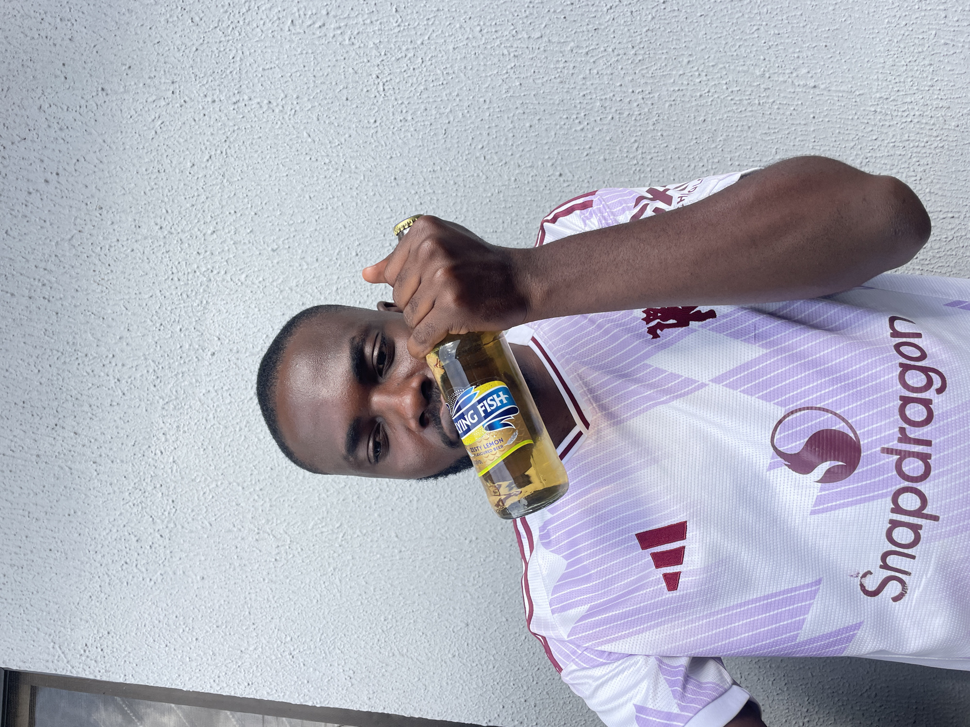 A man with dark skin and a beard is standing outdoors in front of a textured, light-colored wall. He is wearing a white and purple soccer jersey with maroon accents. He is holding a glass bottle of "Flying Fish" flavored beer to his lips and appears to be drinking from it. The bottle has a yellow and blue label. He is also wearing a gold ring on his right index finger. The setting is a wall, which is common in many urban areas, including the city of Jos, Nigeria.