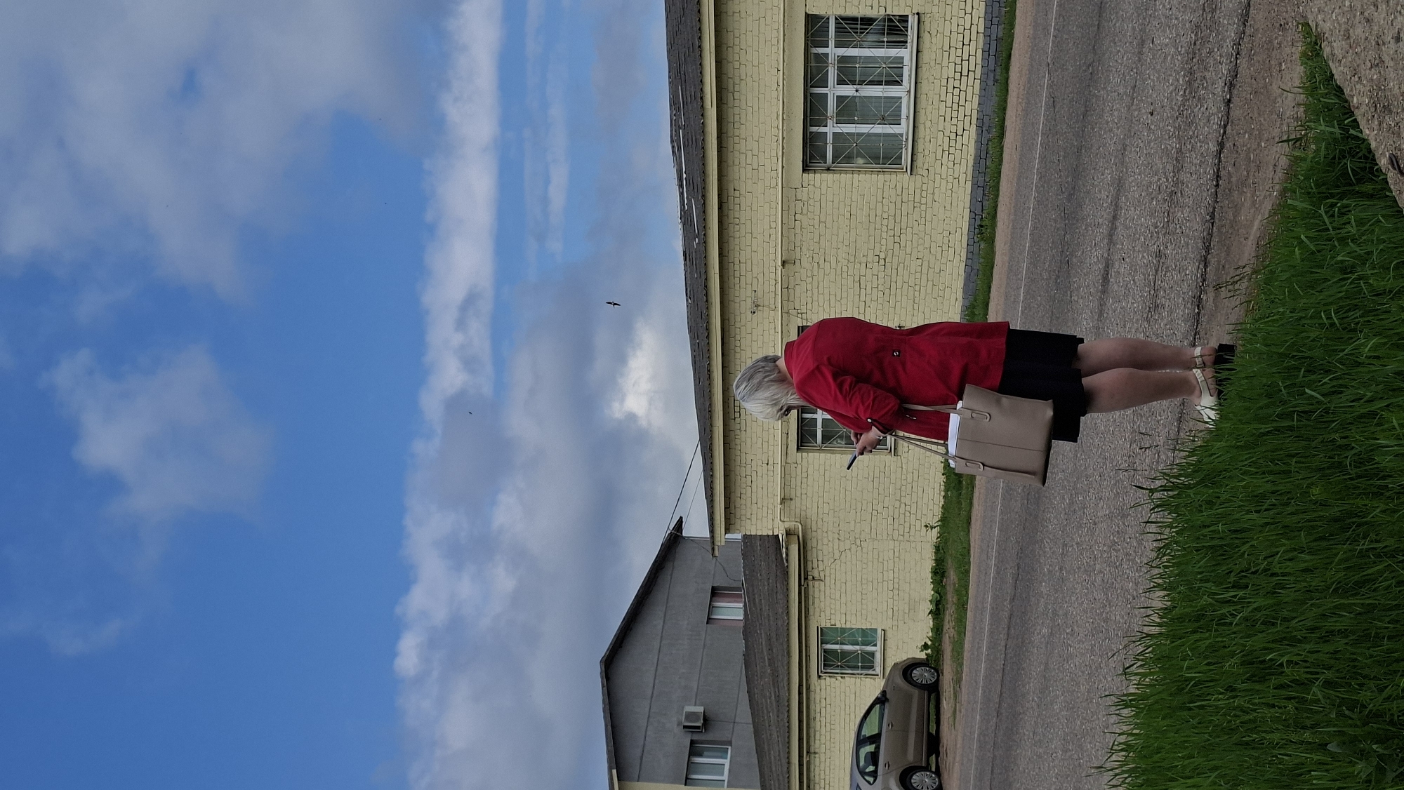 The scene is an outdoor shot in Забалоцкі сельскі Савет, Belarus. An older woman with short gray hair is standing on the side of a paved road. She is wearing a red coat over a black skirt, and has light-colored shoes. She is holding a cell phone in one hand and a beige handbag in the other.
Next to the road, there is a grassy area. Beyond the woman, there is a yellow brick building with white-framed windows. Adjacent to it, there is a gray building with a smaller window. Parked near the gray building, there is a beige car.
The sky above shows a partly cloudy day with blue patches visible among the white clouds.