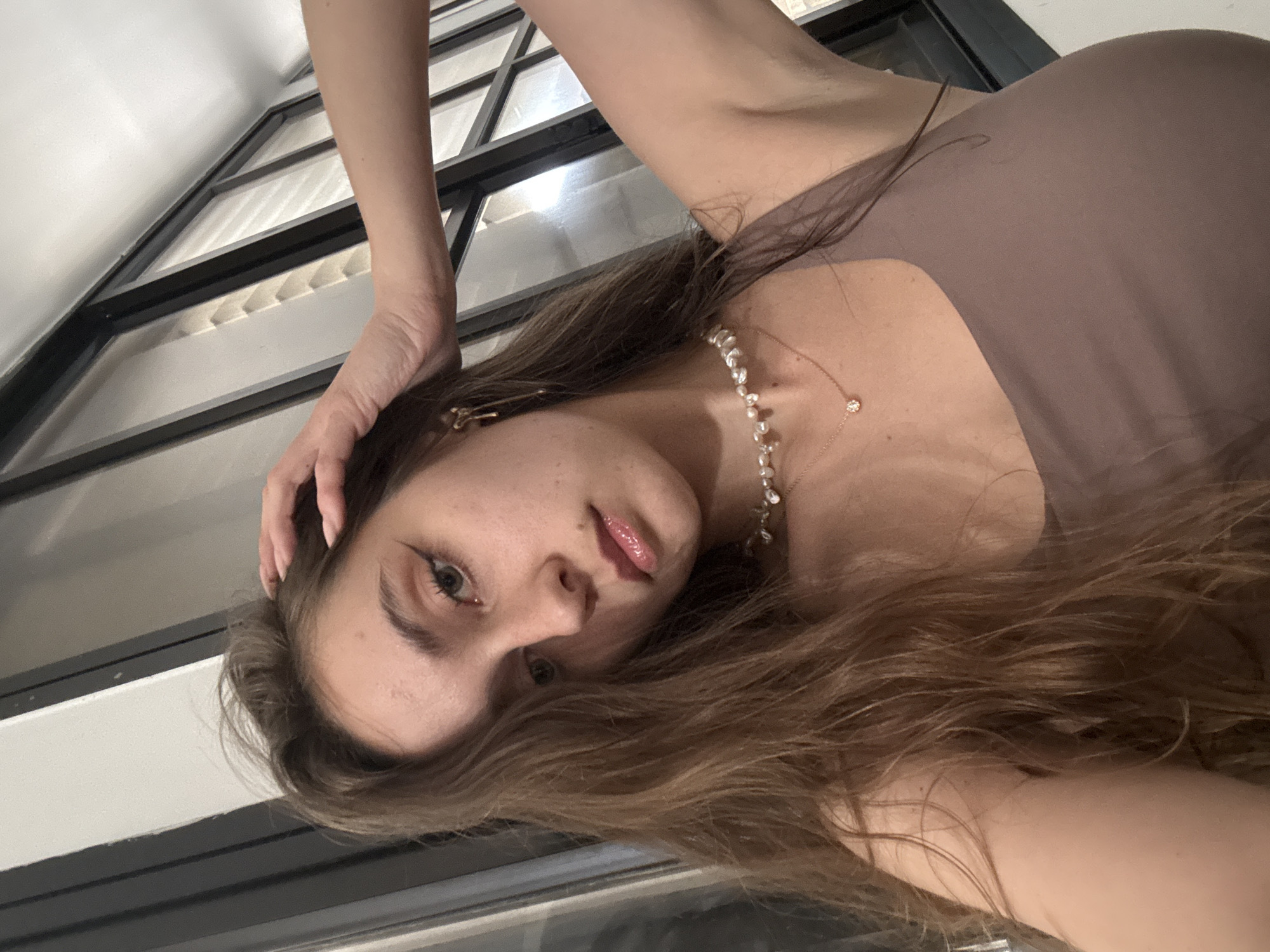 A young woman with long, wavy, light brown hair is positioned in front of a window with a black frame and horizontal white blinds. She is wearing a brown sleeveless garment and a pearl necklace with a small pendant. Her right hand is touching her head. Her face is slightly tilted towards the viewer, and she has light skin, green eyes, and lipstick. The image appears to be taken from a low angle, looking up at the woman. There is no discernible information that indicates the image was taken in Ramat Gan, Israel.