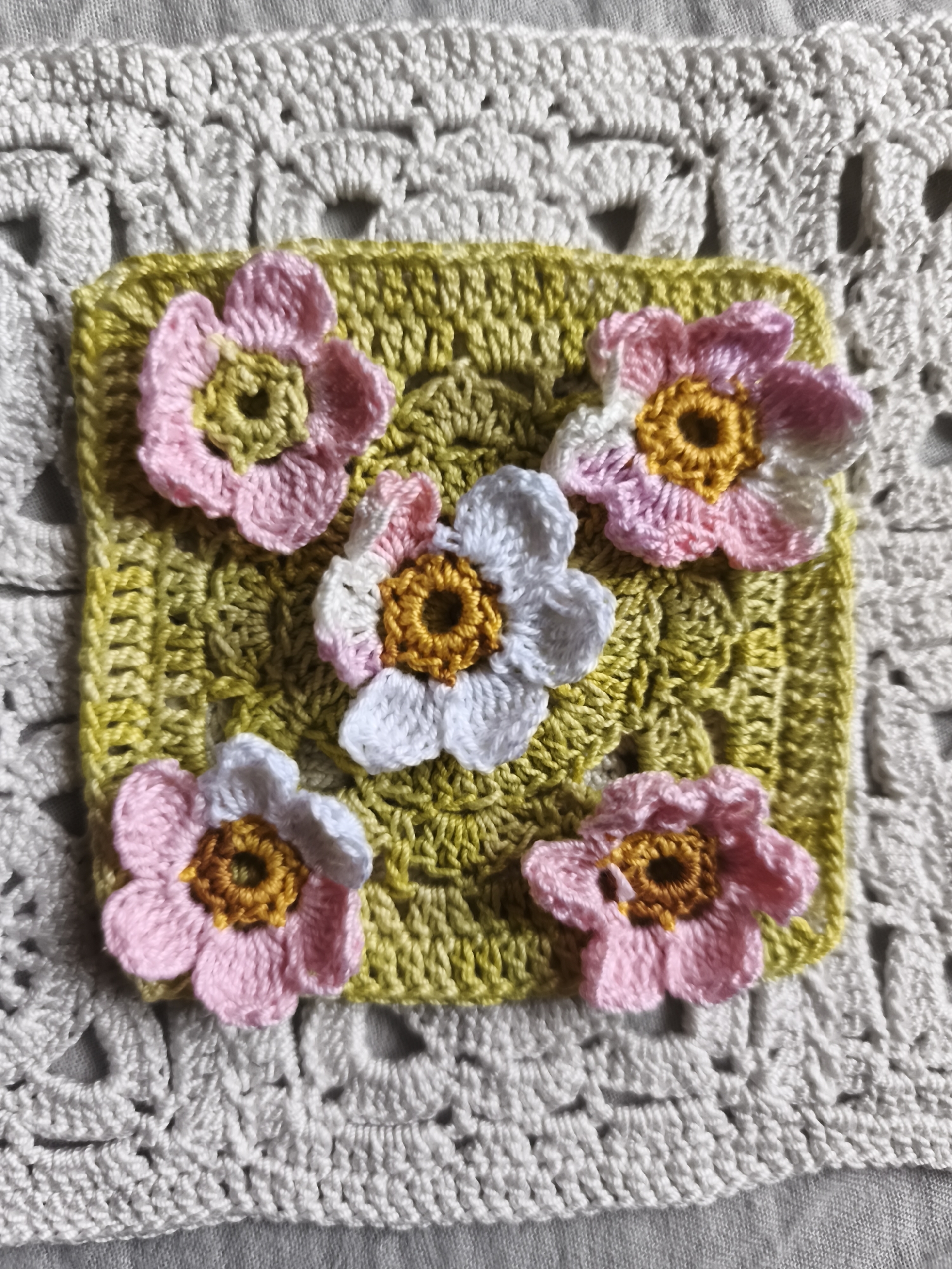 A close-up, overhead shot features a small, square, crochet piece in a light green hue, adorned with five crocheted flowers. Three of the flowers are pink with yellow centers, one is white with a yellow center, and another is white with pink-tipped petals and a yellow center. The green crochet square is placed on top of a larger, white, lacy crochet textile. There are no people or animals present in the image. The scene is indoors, likely on a flat surface such as a table or floor, given the lighting and the texture of the background fabric. The image does not provide any information about the location in the city of Saint-Denis, France.