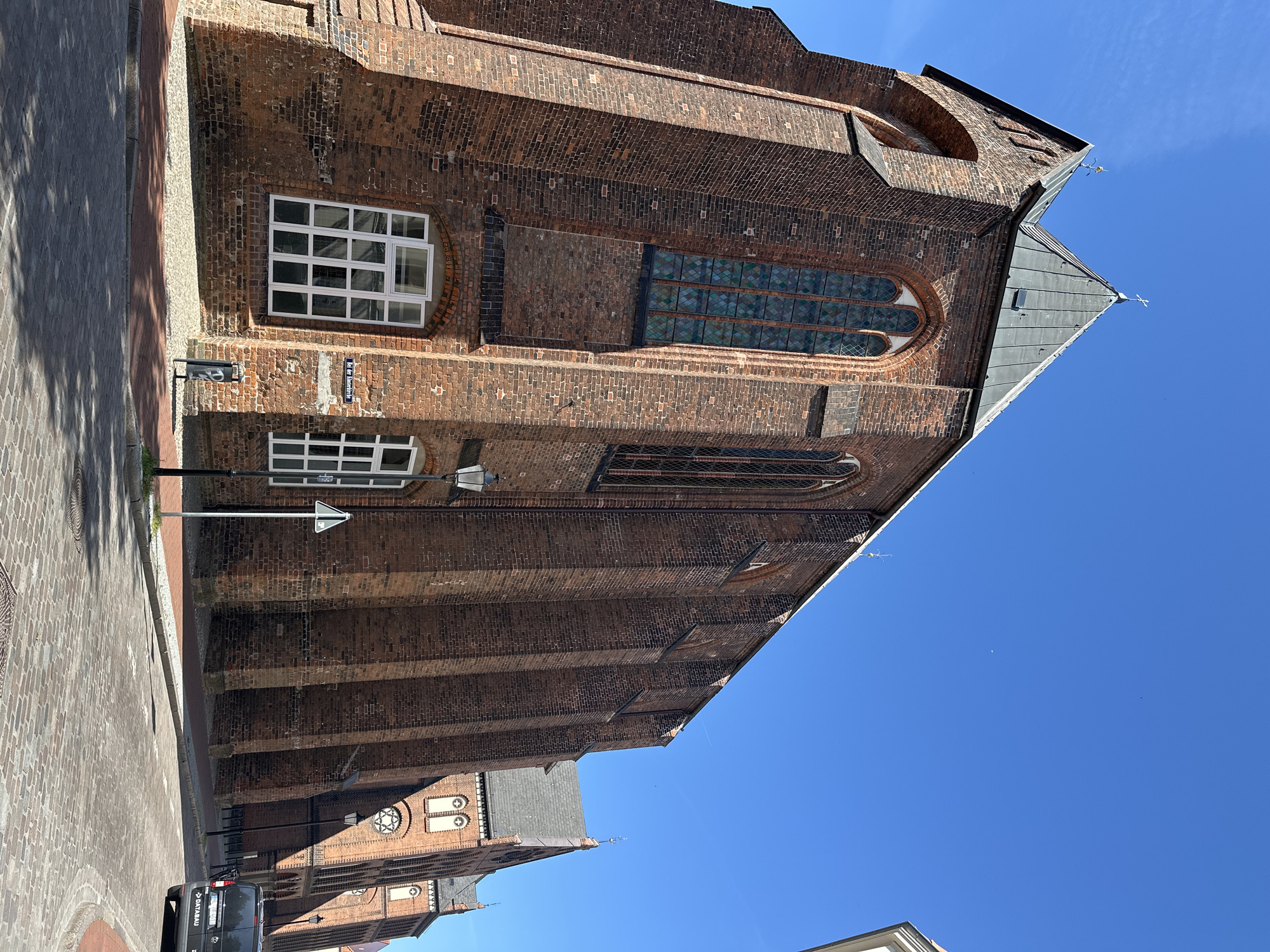 The image shows a street view of the Marienkirche (St. Mary's Church) in Wismar, Germany, on a clear, sunny day. The church is a large, Gothic brick building with a steeply pitched, dark gray roof. Several tall, arched windows with stained glass are visible on its facade. A streetlamp stands in front of the church, and a street sign reading "Bei der Klosterkirche" is mounted on the wall. The street is paved with cobblestones. A dark-colored van with the text "DATABAU" on its rear is parked on the right side of the street. The sky is bright blue with no clouds.