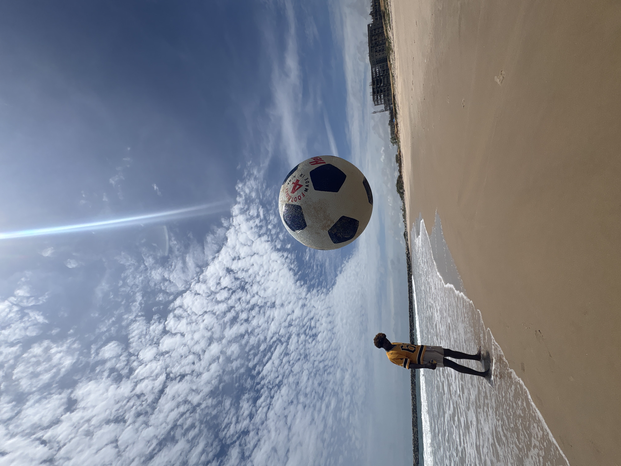 A low-angle, outdoor shot shows a beach scene, with a man standing in the water, a soccer ball seemingly suspended in the air, and the sky filled with clouds. The man is wearing a yellow and black shirt with the number 93 on the back and gray shorts. He is standing at the edge of the water, facing away from the camera. Above him is a soccer ball, appearing to float. The sky is blue with numerous white clouds. A bright, thin band of light is also visible in the sky. The beach extends to the right side of the image. A building under construction is visible in the background. Based on the description, the location is the city of Alaguntan, Nigeria.