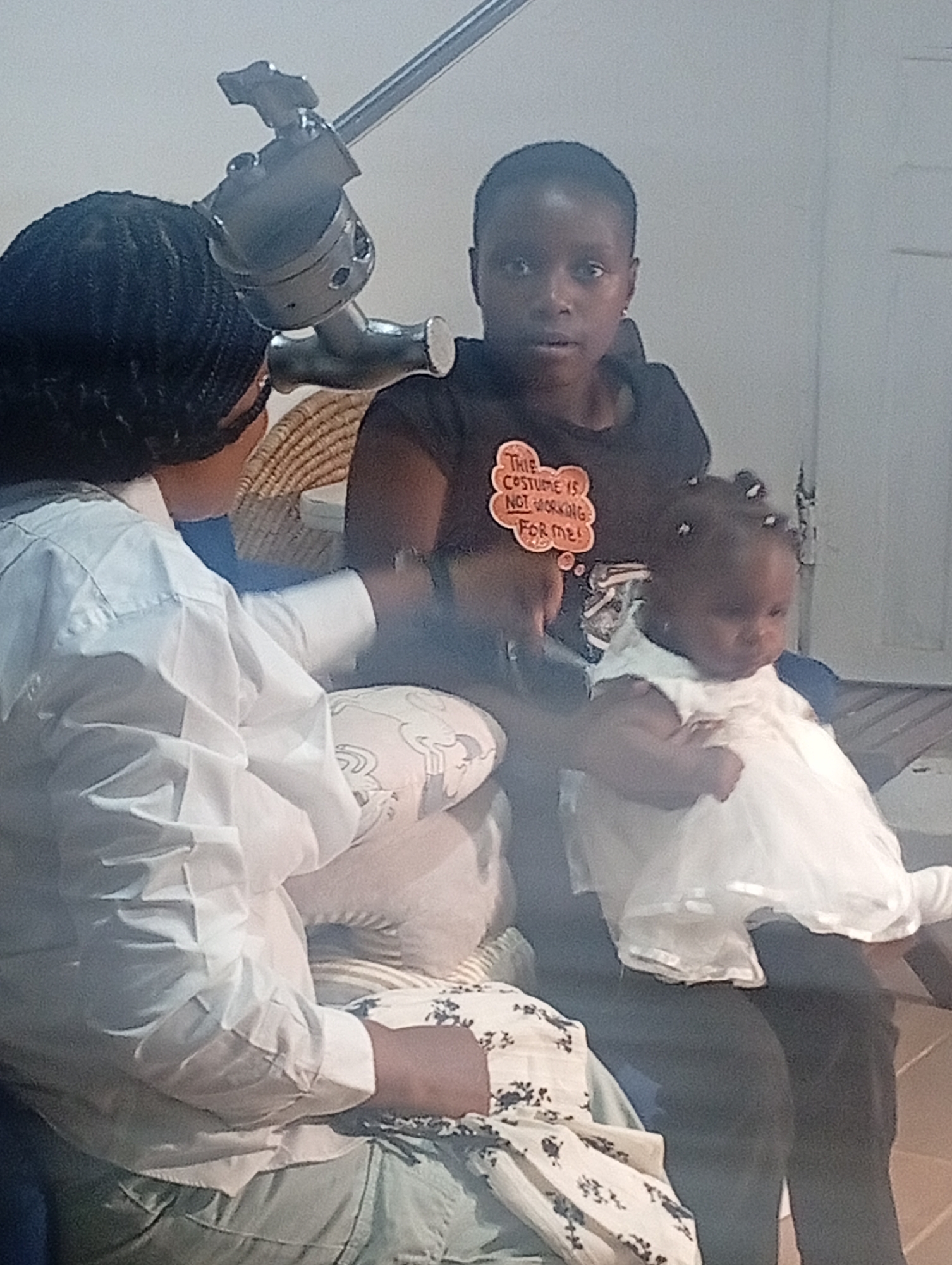 The image depicts three individuals indoors. A woman with braided hair, wearing a white shirt, is seated on a patterned cushion. Another woman, in a dark t-shirt with the text "THIS COSTUME IS NOT WORKING FOR ME!", sits behind her and holds a baby in a white dress. The location is indicated as Barakin Akawo, Nigeria. Behind the seated individuals is a metallic object resembling a lighting or camera stand. The lighting appears to be artificial, illuminating the scene from an unseen source.