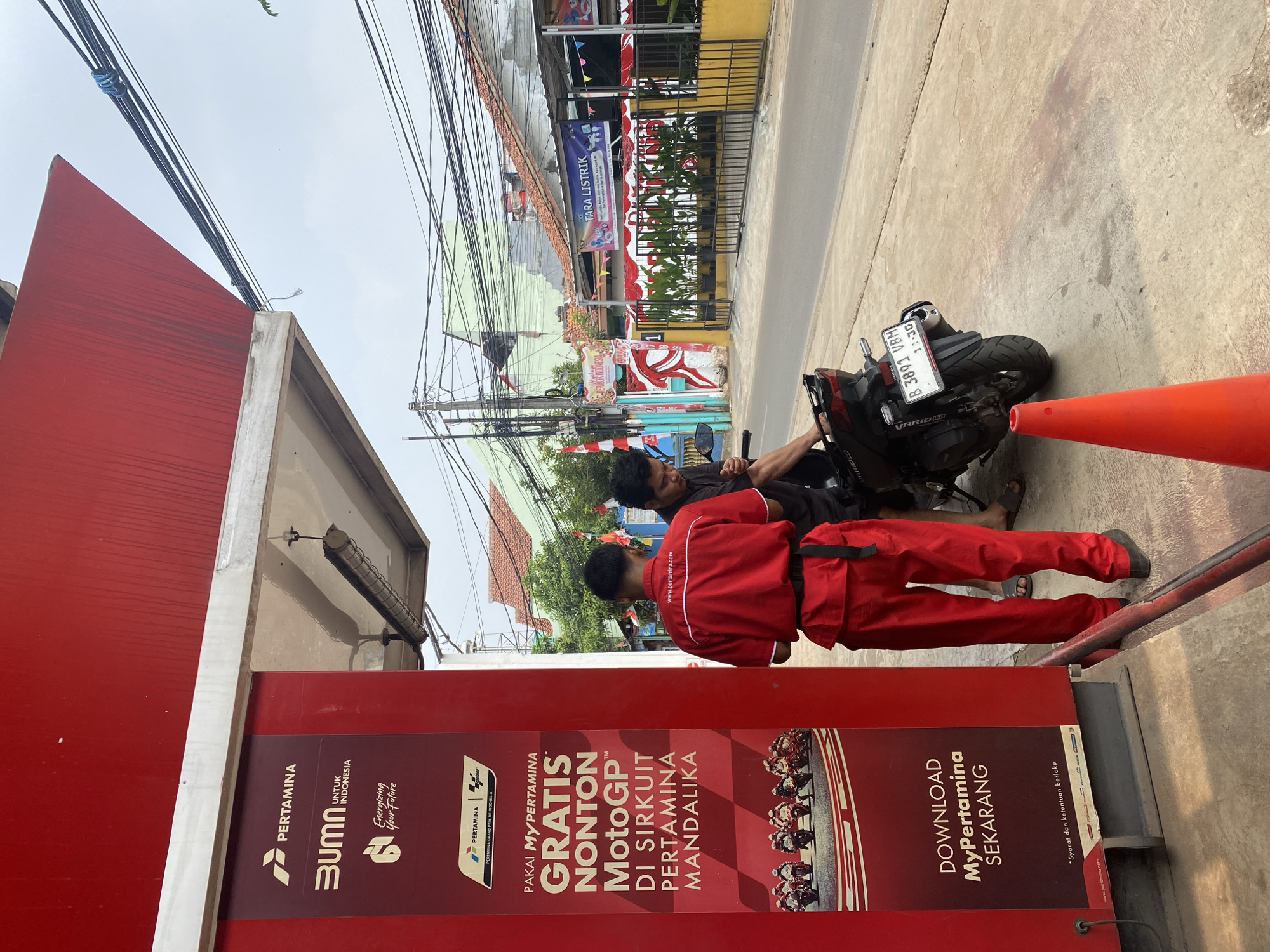 In Tangerang, Indonesia, an outdoor scene unfolds at a Pertamina gas station. Two men are present. One man, wearing a red Pertamina uniform consisting of a shirt and pants, stands facing a red kiosk with Pertamina branding. The other man, in a dark shirt, is partially obscured, leaning over a black motorcycle parked on the sidewalk. The motorcycle has a license plate that reads "B 3893 VBM". The kiosk displays advertisements for Pertamina, including a promotion for free MotoGP viewing at the Pertamina Mandalika Circuit and a call to download the "MyPertamina" app. In the background, a street is visible with overhead electrical wires, buildings with red tile roofs, and a fence with potted plants. A bright orange traffic cone stands on the concrete sidewalk in the foreground. The weather appears overcast.