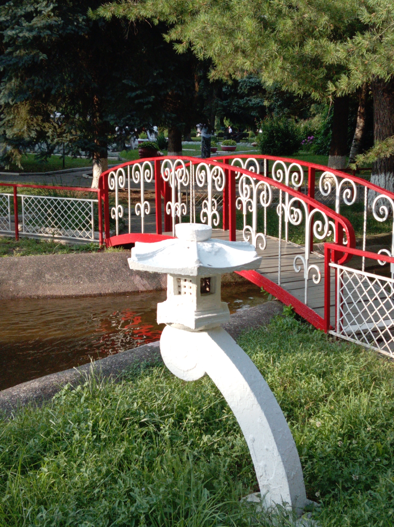 The image captures a serene park scene, likely in Yessentuki, Russia, with a picturesque red and white arched bridge as the central focus. The bridge, adorned with decorative white scrollwork, spans a narrow, dark body of water. To the left of the bridge, a white wire fence encloses a patch of grass. In the foreground, a white, pagoda-style garden lamp with a square base and tiered roof stands beside the water, partially obscuring the bridge's walkway. Lush green grass covers the ground around the lamp and the water's edge. Behind the bridge, a variety of trees, including dark green conifers and lighter green deciduous trees, create a verdant backdrop, with sunlight filtering through their branches. The sky is not visible.