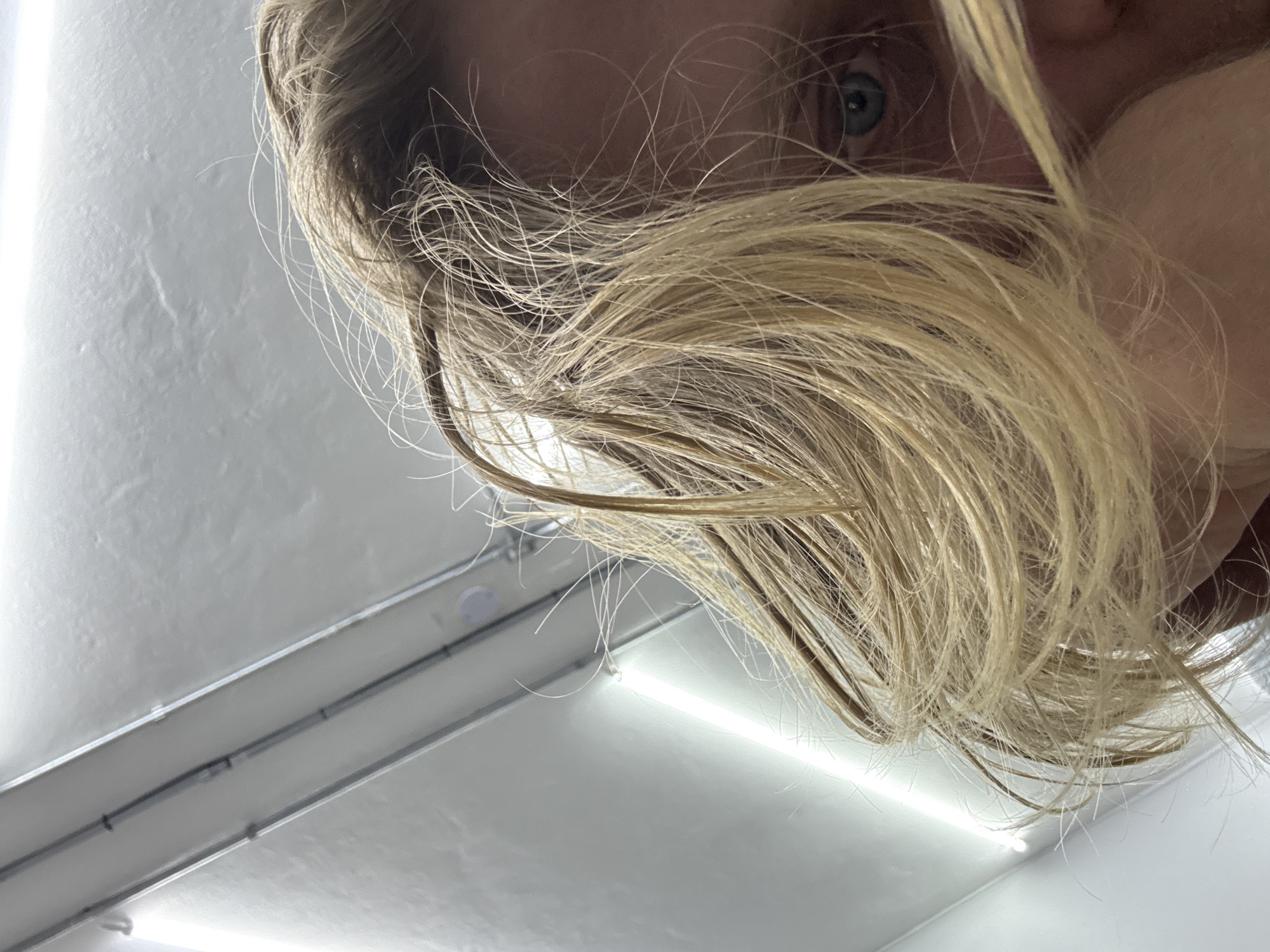 The image shows a close-up, low-angle shot of a person's face, partially obscured by their blonde hair. The person's eye, with a blue iris, is visible on the right side of the frame. The background is a white ceiling with fluorescent light fixtures and some metallic conduit. The scene appears to be indoors, possibly in a bathroom or a similar utilitarian space, given the lighting and ceiling design. There is no indication of the specific location within São Paulo, Brazil.