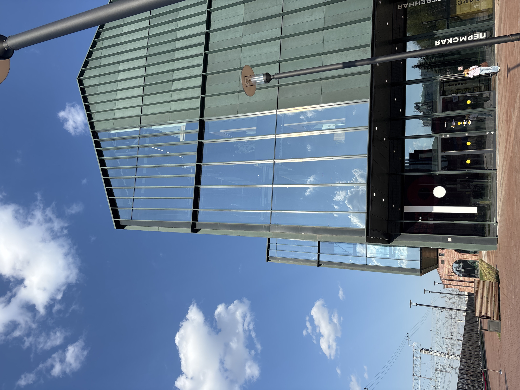 The image shows a modern glass and metal building under a partly cloudy blue sky. The building's glass reflects the sky and clouds. A black lamp post is attached to the building. The building has signage in Russian: "Пермская". A person in pink is visible inside. Below the building are electrical lines, and in the distance is a red brick building. The setting is likely a station or transportation hub in Perm, Russia.