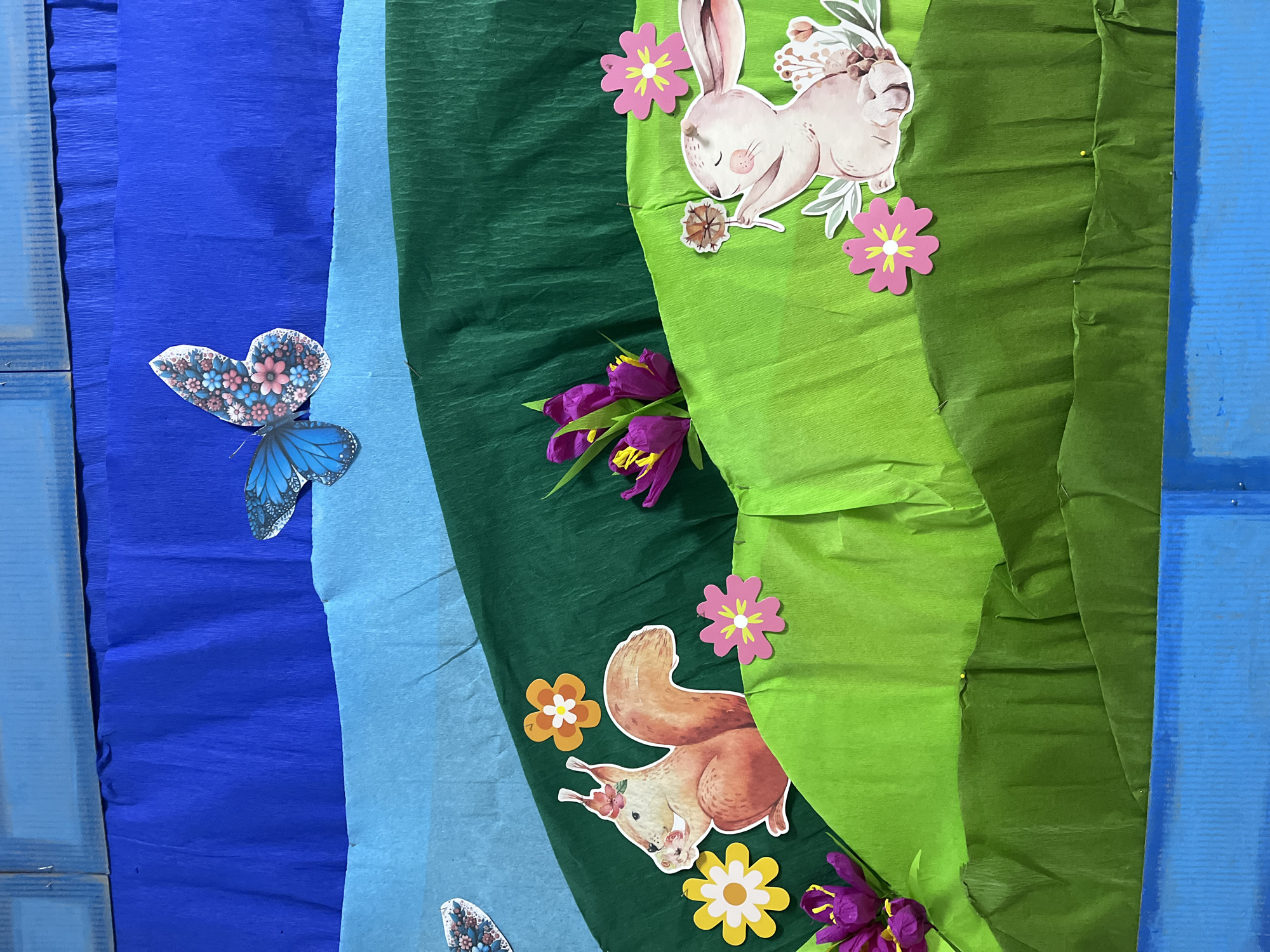 The image shows a backdrop or wall decoration with vertical stripes of blue, light blue, dark green, and light green crepe paper. The blue and light blue stripes are on the left side of the image, while the dark green and light green stripes are on the right.
Adhered to the crepe paper are several cut-out images: a butterfly on the blue stripe, a squirrel and several flowers on the dark green stripe, and a rabbit and more flowers on the light green stripe. There are also some purple flowers attached to the dark green and light green stripes. The background appears to be made of blue painted wooden planks.