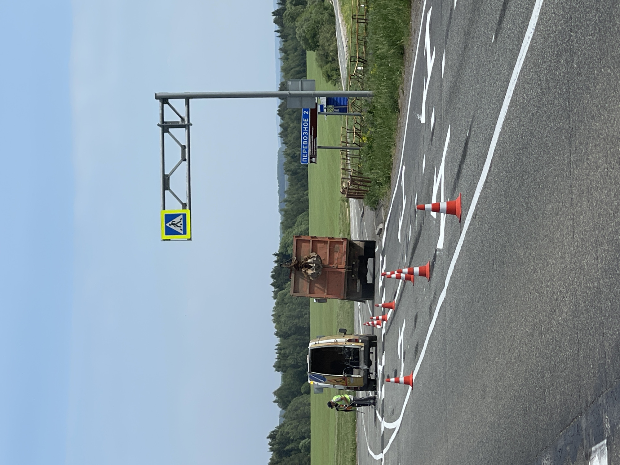 The image depicts a road in Перевозное, Russia. A truck and a van are parked on the side of the road, near a worker who appears to be performing road maintenance. Orange and white traffic cones are set out in a line along the edge of the road. A pedestrian crossing sign and a sign indicating "Перевозное 2" are visible. The surrounding environment consists of a grassy field and trees. The sky is clear and blue.
