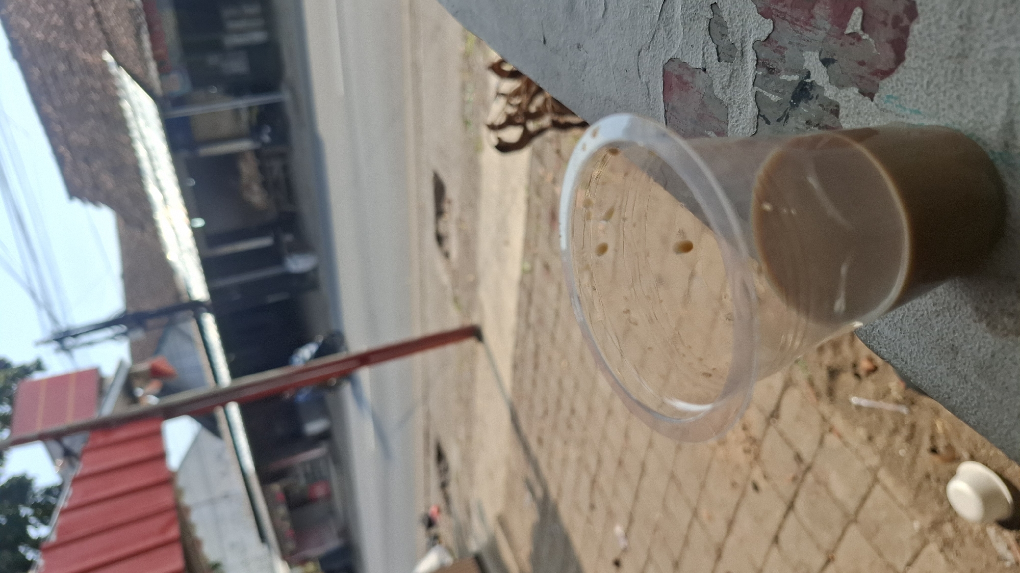 The image shows a close-up view of a clear plastic cup filled with a light brown beverage, likely coffee or tea, placed on a textured, gray surface. The cup has some condensation and a few small bubbles or imperfections visible inside and on its surface. Behind the cup, a street scene is visible with buildings, a road, and some foliage. A red pole is prominent in the background, possibly part of a structure or signage. The street appears to be in an urban or semi-urban area, with storefronts lining one side of the road. The overall lighting suggests it is daytime, with shadows indicating sunlight. The perspective is low, looking up at the cup and then out towards the street. No people are clearly identifiable. The image could have been taken in Cilaku, Indonesia, given the architectural style of the buildings and the presence of what appears to be street vendors or small shops.
