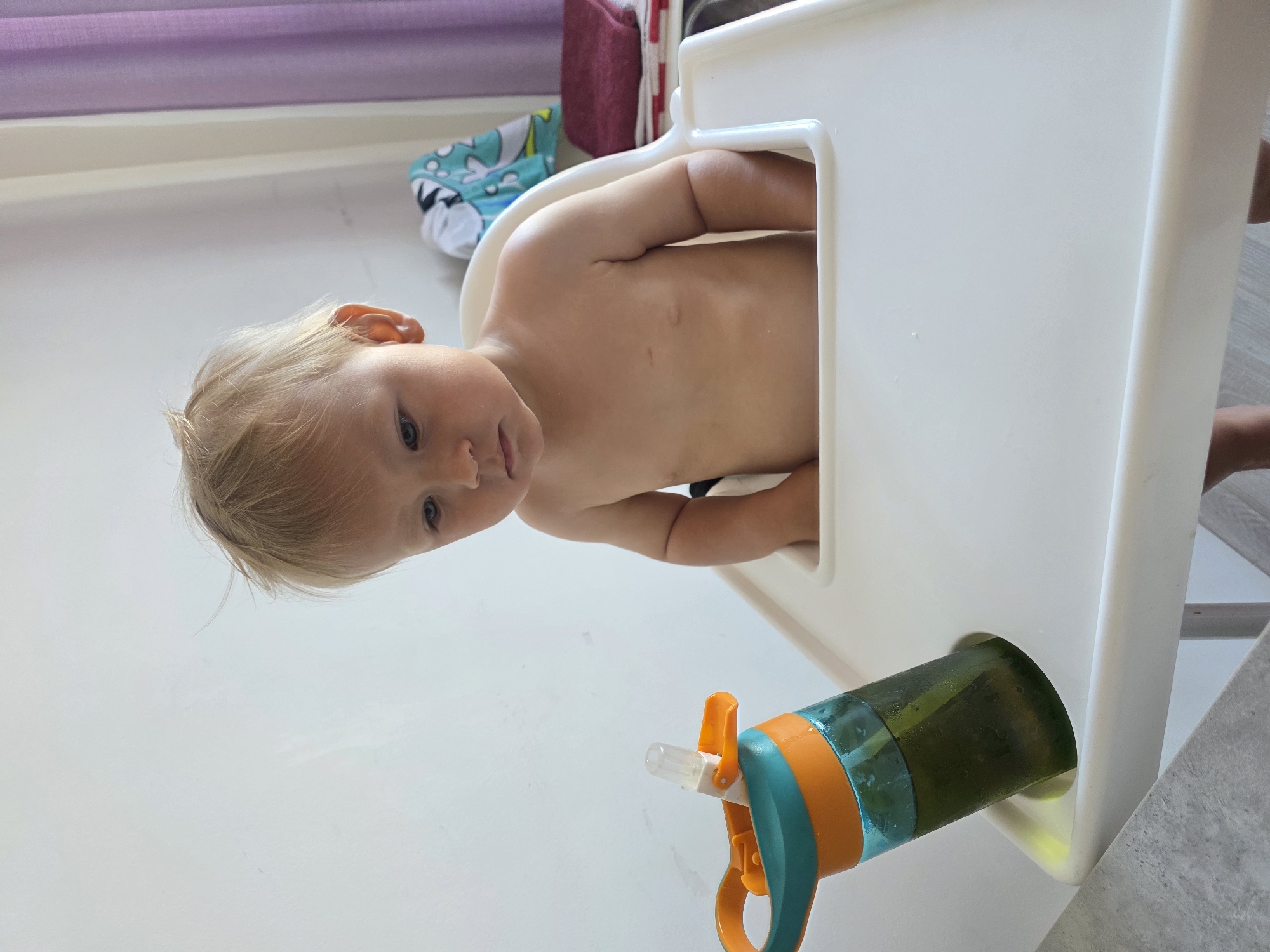 A young child with blonde hair and blue eyes is sitting in a white high chair. The child is facing forward, looking towards the camera with a curious expression. Their upper body is exposed, showing bare shoulders and a pale torso. The child's hands are visible, resting on the tray of the high chair.
In front of the high chair, a green and orange water bottle with a straw is placed on the floor. The bottle appears to be partially filled with a green liquid. The high chair is positioned in a brightly lit room, possibly a dining area or kitchen, with a light-colored floor and a textured grey surface visible to the right. In the background, a portion of a window with purple curtains is visible, suggesting it is daytime. A patterned fabric, possibly a bib or towel, with a shark design in blue and white, is also visible behind the child. The overall scene is casual and domestic, capturing a moment of a child in their high chair. There is no visible text in the image. The setting appears to be indoors. The lighting suggests it is daytime.