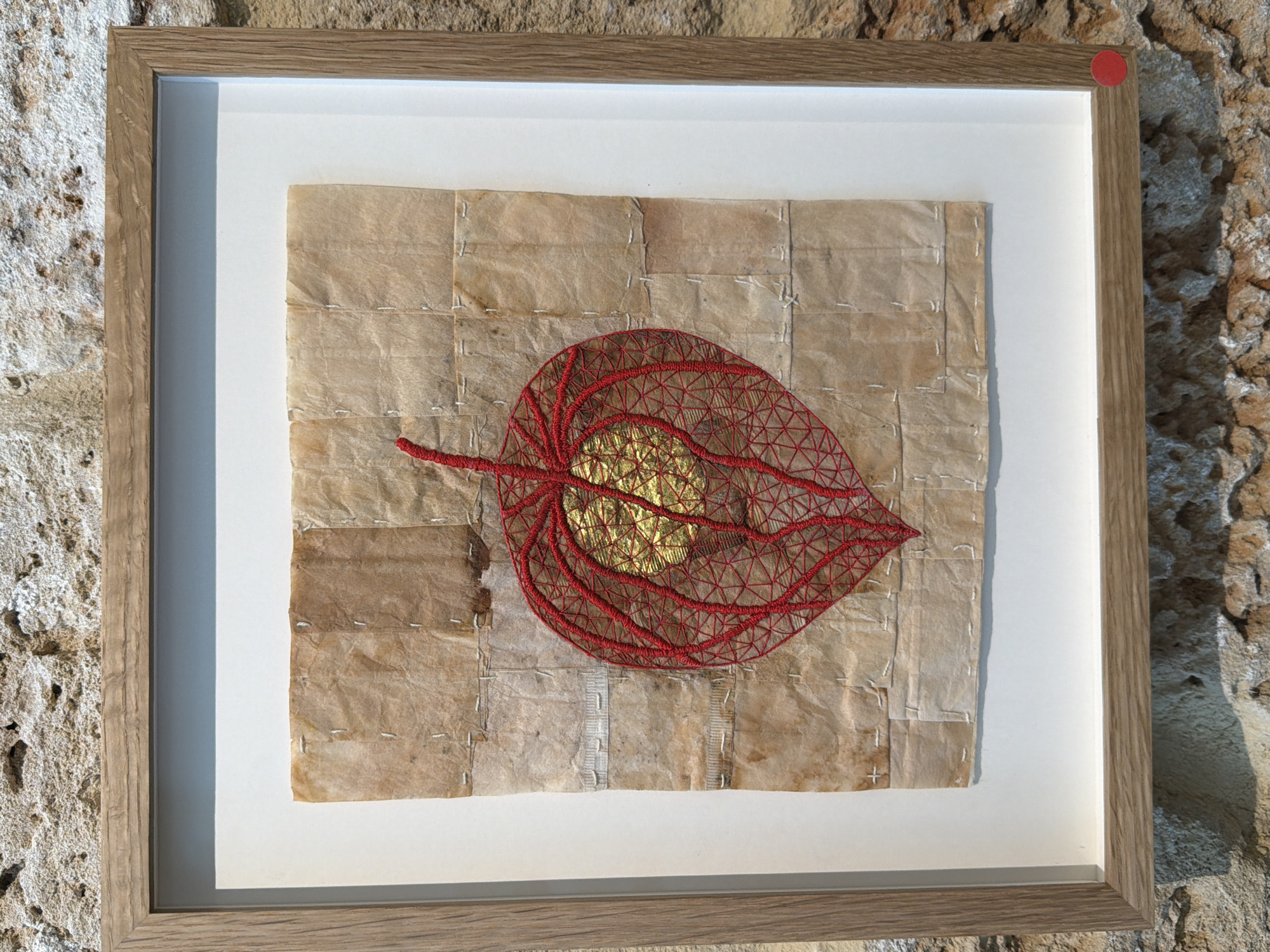 A framed mixed-media artwork is displayed against a rugged, light-colored stone wall, suggesting an exhibition space or a unique home interior, possibly in Tel-Aviv, Israel. The artwork is housed within a light brown wooden frame with a white mat board.
The central piece features a delicate, intricate representation of a red, skeletal-like leaf or seed pod, reminiscent of a Chinese lantern (Physalis alkekengi). This red structure appears to be crafted from fine thread or wire, forming a web-like pattern that highlights the leaf's veins. At the heart of this red form, a crumpled, shimmering golden object or material provides a vibrant focal point.
The background of the artwork consists of numerous small, rectangular pieces of aged, light brown, textured material, possibly paper or thin fabric, stitched together with white thread. These stitches are visible, creating a grid-like, patchwork texture behind the red leaf.
A small, circular red sticker is affixed to the top right corner of the wooden frame. The scene is brightly illuminated by natural light, indicating it is daytime. There is no visible text in the image.