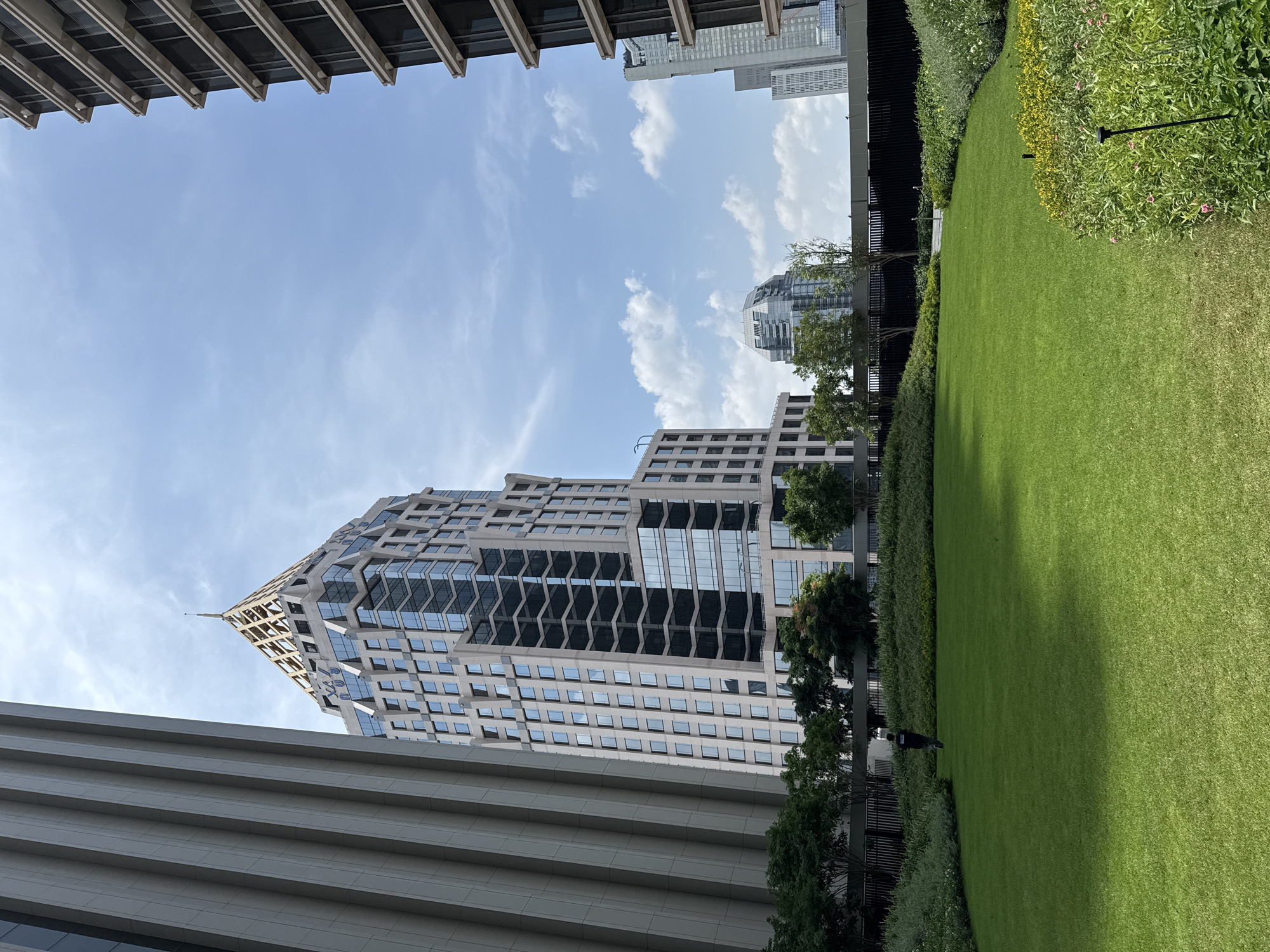 The image captures a scene in Bangkok, Thailand, showcasing the city's modern architecture and urban landscaping. The focal point is a tall, contemporary building with a unique, textured facade, which appears to be a high-rise office or residential tower. Below, the image includes a manicured lawn with lush greenery. A person is walking along a path bordering the lawn and the building. The sky is partly cloudy. A building featuring horizontal lined architecture is present. The perspective is from below, looking upwards, creating a sense of scale and highlighting the contrast between the natural and built environments.