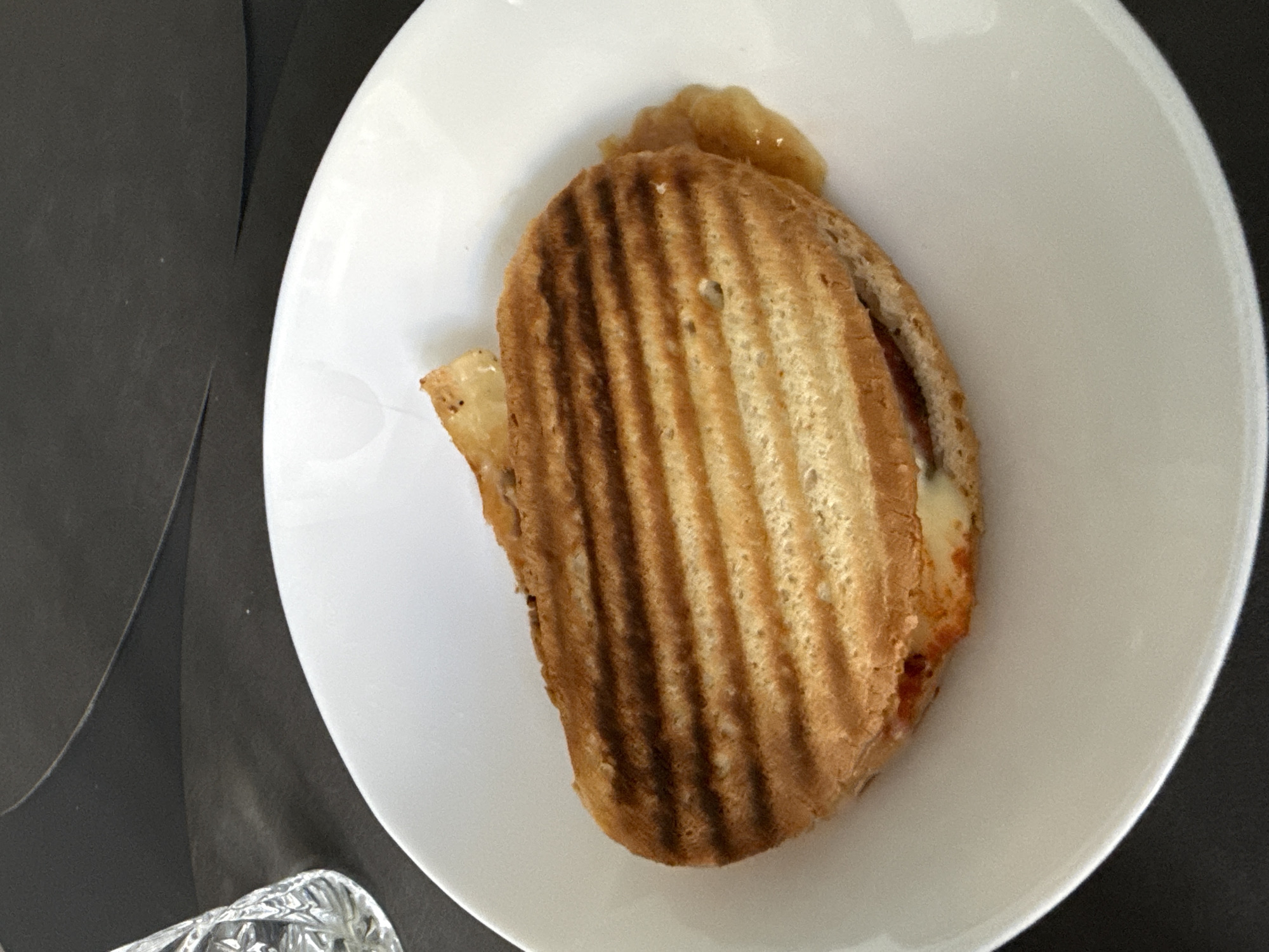 A close-up overhead shot shows a grilled panini sandwich with visible grill marks on a white plate. A small amount of what appears to be apple sauce is on the side of the panini. The sandwich is partially open, revealing cheese and possibly tomato sauce inside. The plate is set on a dark, textured surface. To the left of the plate, a portion of a clear, faceted glass is visible, suggesting the presence of a drink. The background is dark and out of focus. There are no people or animals present in the image. The scene is indoors, likely a restaurant or home setting. The location in Stara Zagora, Bulgaria is not discernible from the image itself.