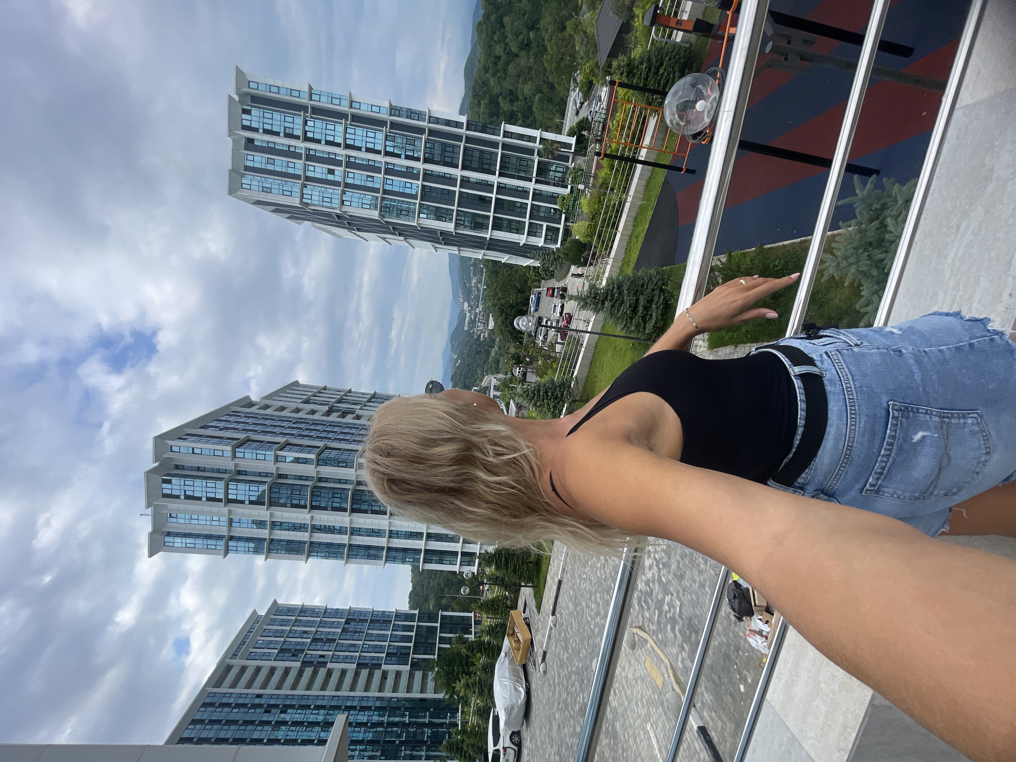 The image shows a blonde woman standing on a balcony, looking out at a cityscape. She is wearing a black tank top and denim shorts. In the background, there are several modern apartment buildings with blue glass windows. The buildings are surrounded by lush green trees and rolling hills, suggesting a location in a city with a lot of natural scenery. The sky is cloudy. The woman is holding her phone up as if taking a selfie, with her arm extended to the left. There is a children's playground visible on the ground below the balcony. Based on the architectural style of the buildings and the surrounding landscape, the location is likely in Sochi, Russia, possibly in the Adler district, known for its contemporary residential complexes and proximity to the Caucasus Mountains.
