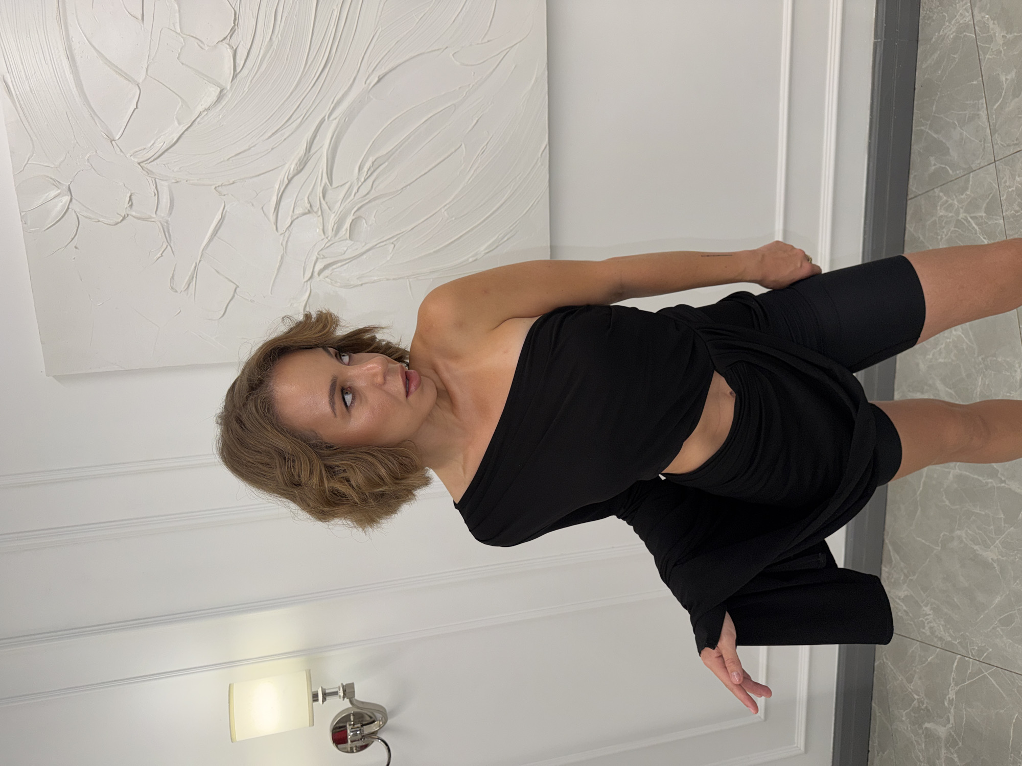 A woman stands indoors, facing right, her gaze directed away from the camera. She has medium-length, wavy brown hair and is wearing a black one-shoulder top with cutouts at the waist, paired with black biker shorts. Her left arm is bent, with her hand resting on the draped fabric of her top. The background features a white wall with vertical paneling and a modern, textured white artwork. A wall-mounted sconce with a white shade is visible on the left. The floor is tiled with a marble pattern. The setting appears to be a studio or a well-lit interior space. There is no indication of the location being Moscow, Russia.