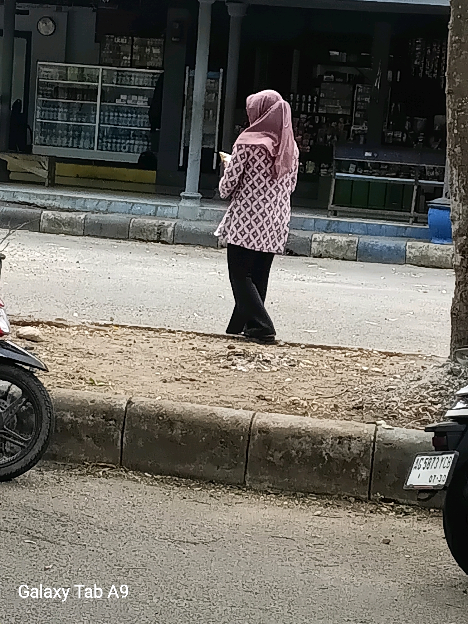 The image captures a street scene in Trenggalek, Indonesia. A woman wearing a pink hijab and a patterned tunic is walking away from the camera, holding a mobile device. She is dressed in black pants and dark shoes. To her left, the front wheel and fender of a black motorcycle are visible. The background consists of a building with glass display cases filled with merchandise, and a sidewalk with a concrete curb. A tree trunk is prominent on the right side of the frame. A license plate on a motorcycle, partially visible on the lower right, reads "AG 5873 YCB 07-30". The ground is paved with asphalt and has scattered debris. The text "Galaxy Tab A9" is overlaid on the lower left corner of the image.