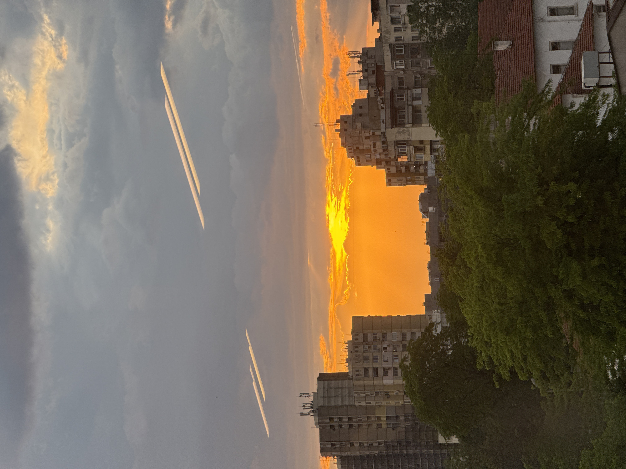 The image shows an urban landscape at sunset. Buildings of varying heights, mostly residential apartments, are visible in the center. The sunset is a vibrant orange, illuminating the sky behind the buildings and partially obscured by clouds. Tree foliage is seen on the right side, partially obscuring some of the buildings. Jet trails are present in the upper left portion of the sky. No people are visible. It is not possible to determine if the photo was taken in Plovdiv, Bulgaria.