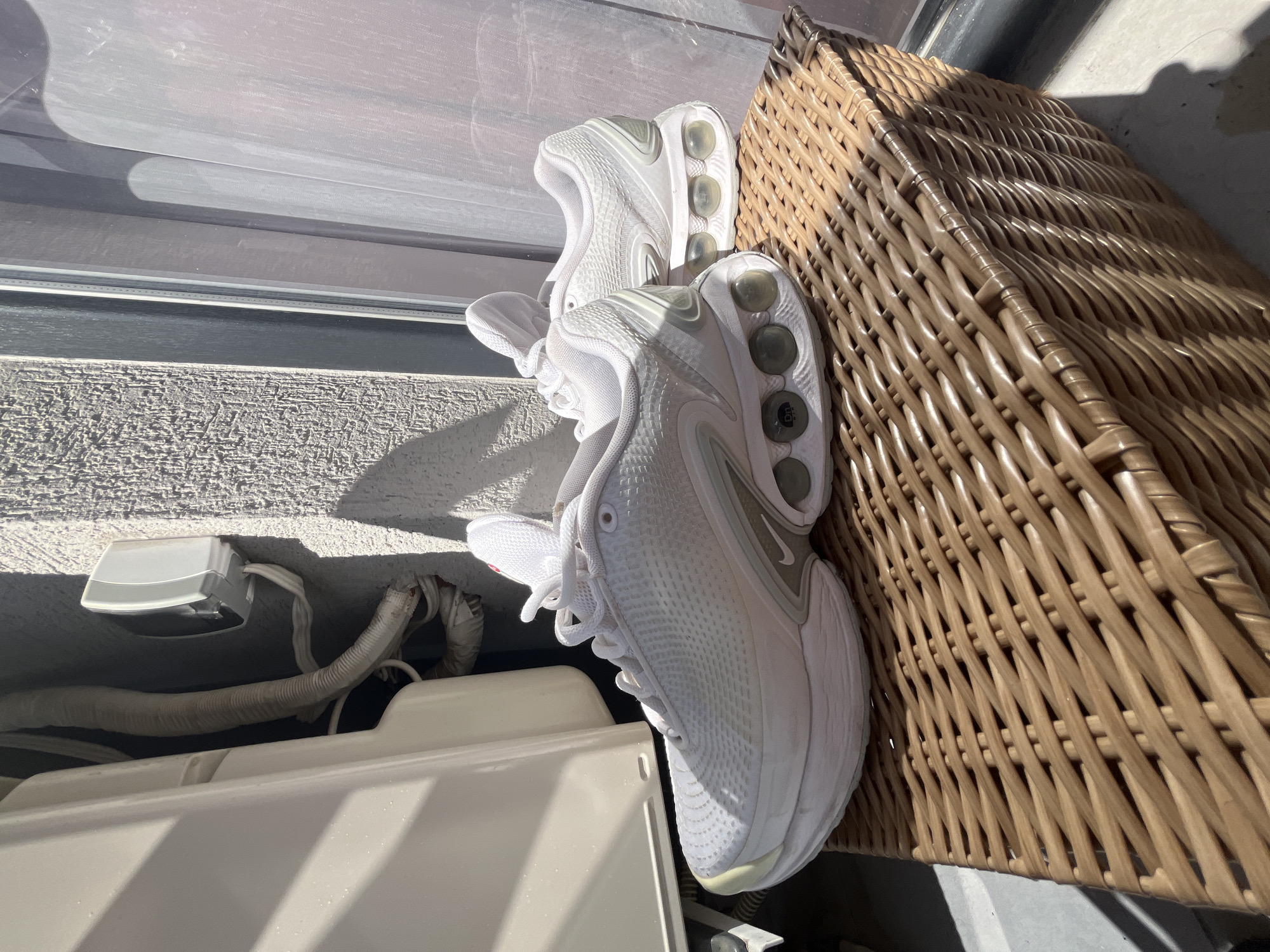 The image shows a pair of white athletic shoes resting on a woven basket. The shoes are positioned vertically, with their soles facing upwards. The basket appears to be made of natural fibers and has a warm, brown tone. The scene is outdoors, indicated by the strong sunlight casting shadows on the ground and the woven basket. The sunlight suggests it is daytime, likely late morning or afternoon. The background includes a textured gray wall and some white piping and a white electrical box, which could be part of an outdoor air conditioning unit or similar apparatus. The setting appears to be a residential or commercial outdoor space. There is no visible text in the image. The overall impression is one of simple domesticity or a moment of casual outdoor living.