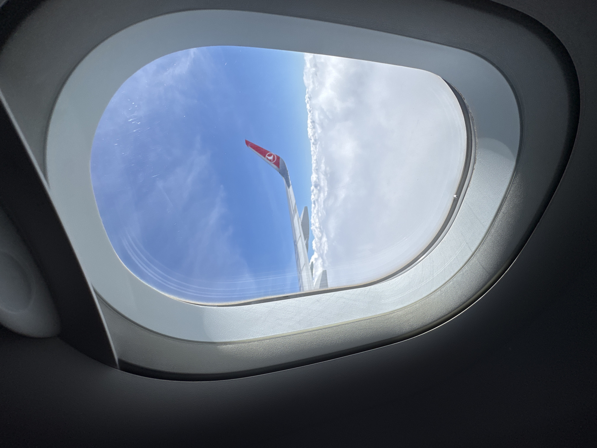The image depicts a view from an airplane window, showcasing the aircraft's wing extending into a blue sky on one side and a dense cloud cover on the other. The wingtip is painted red with the Turkish Airlines logo. The window frame is clean and bright, with the interior of the plane dimly lit. The location is not possible to determine from this picture alone.
