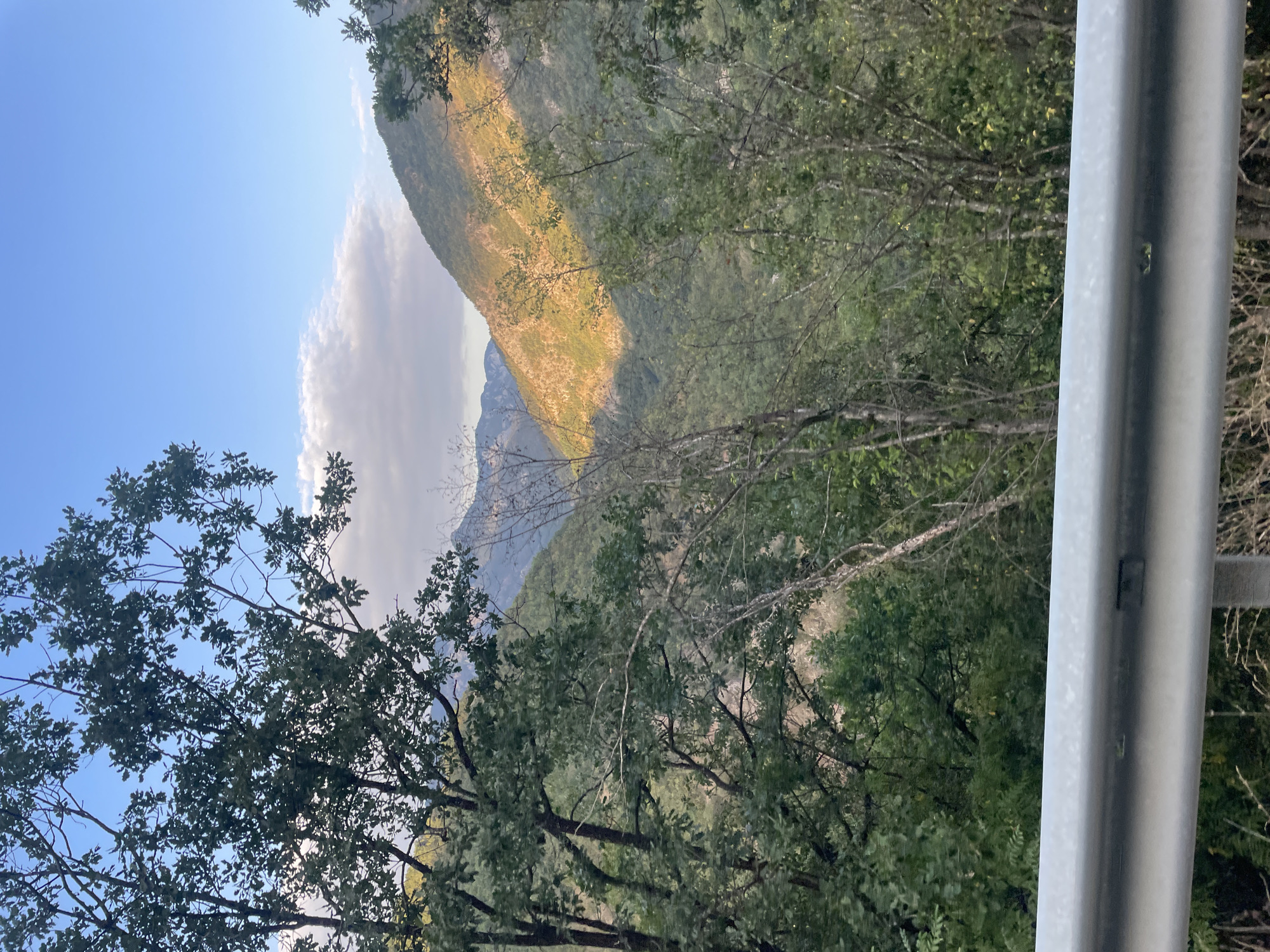 A wide-angle shot captures a sunlit mountain valley with a clear blue sky and scattered clouds. The foreground is dominated by a metal guardrail, partially obscuring dense green foliage and trees. The midground features a valley filled with lush green trees and vegetation. In the background, a mountain slope is illuminated by the warm golden light of the sun, casting dramatic shadows. The scene appears to be located in the city of Yugovo, Bulgaria. No people are visible in the image.