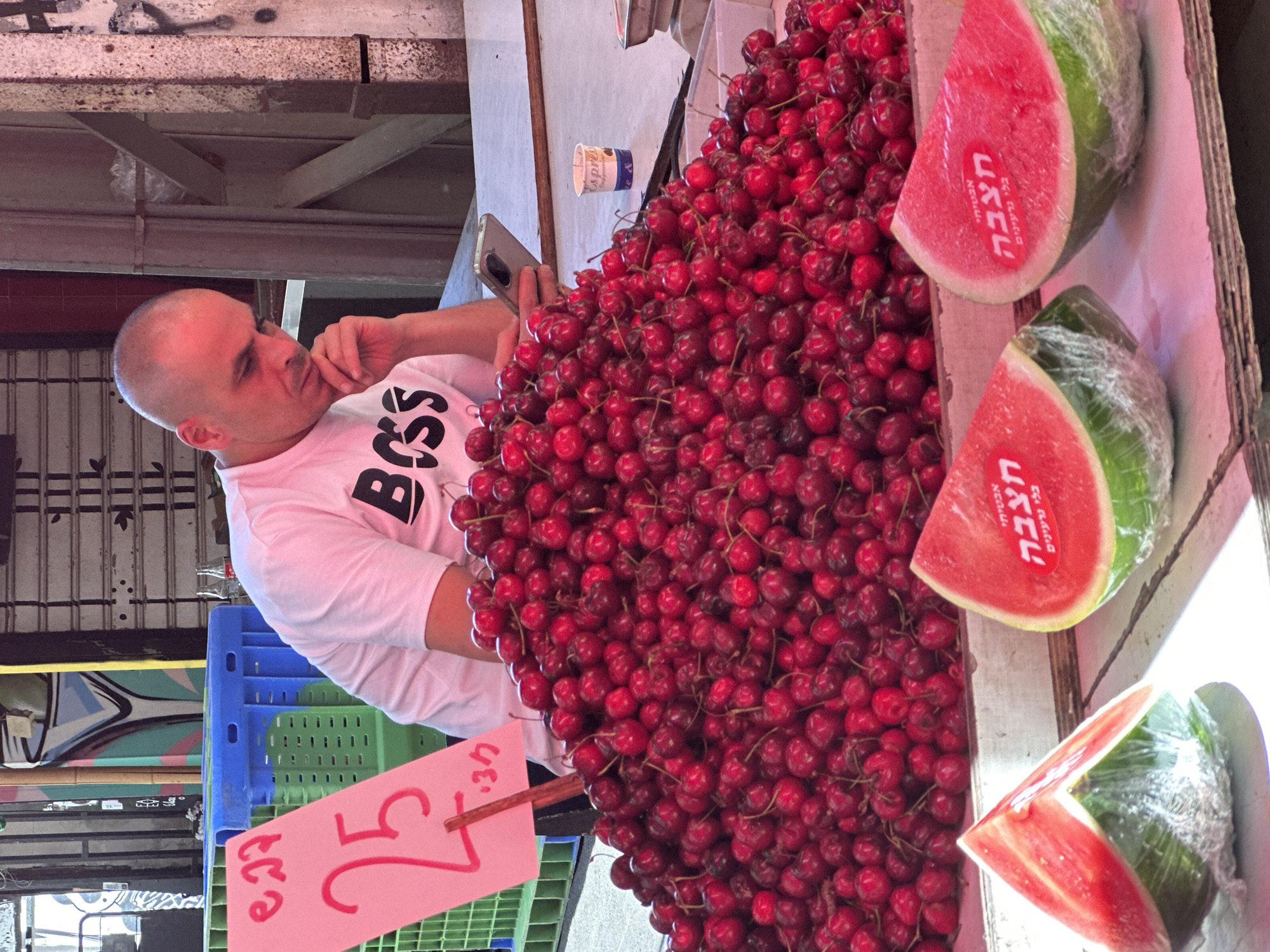 This image captures a market stall in Tel-Aviv, Israel, featuring a vendor and various fresh produce. A bald adult male, wearing a white t-shirt with "BOSS" printed in black, is seated behind a large pile of cherries. He is engrossed in his smartphone, held in his right hand, with his left hand supporting his chin. His gaze is directed downwards towards the phone.
In the foreground, a substantial mound of dark red cherries, many with stems, dominates the display surface. To the lower right, three slices of watermelon, wrapped in clear plastic film, are visible. Two of these watermelon slices bear red stickers with white Hebrew text reading "אבטיחי חצבה בלאי גרעינים" (Hatzva Watermelons, Seedless).
To the left of the cherry pile, a pink rectangular sign is positioned, displaying handwritten red Hebrew text and numbers. The prominent large number "25" is visible, likely indicating a price, with smaller text "₪ 27" and "ל/ק 30" also present. On the table to the right of the vendor, a small white paper cup with "expres... cappucino" is situated near a partially visible metal scale.
The background reveals a typical market setting with closed metal roll-down shutters, various metal structural elements, and a colorful mural or graffiti on a wall. Blue and green plastic crates are also visible behind the pricing sign. The scene is illuminated by natural light, indicating an outdoor or semi-outdoor location.