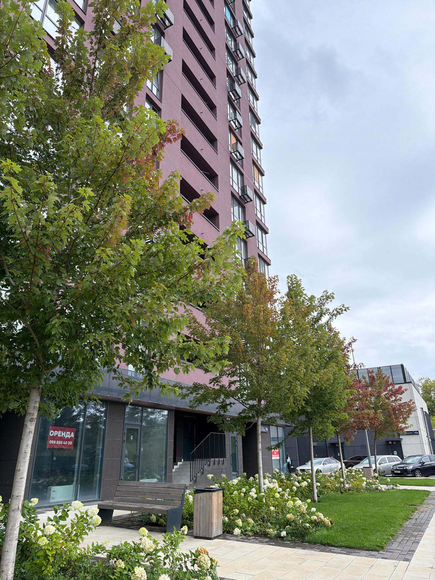 The image captures a street view in Kyiv, Ukraine, showcasing a modern multi-story residential building with a maroon facade. In the foreground, several trees with green and autumn-colored leaves are positioned along a paved walkway. A wooden bench and a trash receptacle are situated near the building's entrance, which features large glass windows. A red sign with the word "ОРЕНДА" (which translates to "RENT" in Ukrainian) and a phone number is visible on one of the windows. Several cars are parked along the street, and the sky is overcast. The surrounding area is landscaped with grass and flowering bushes.