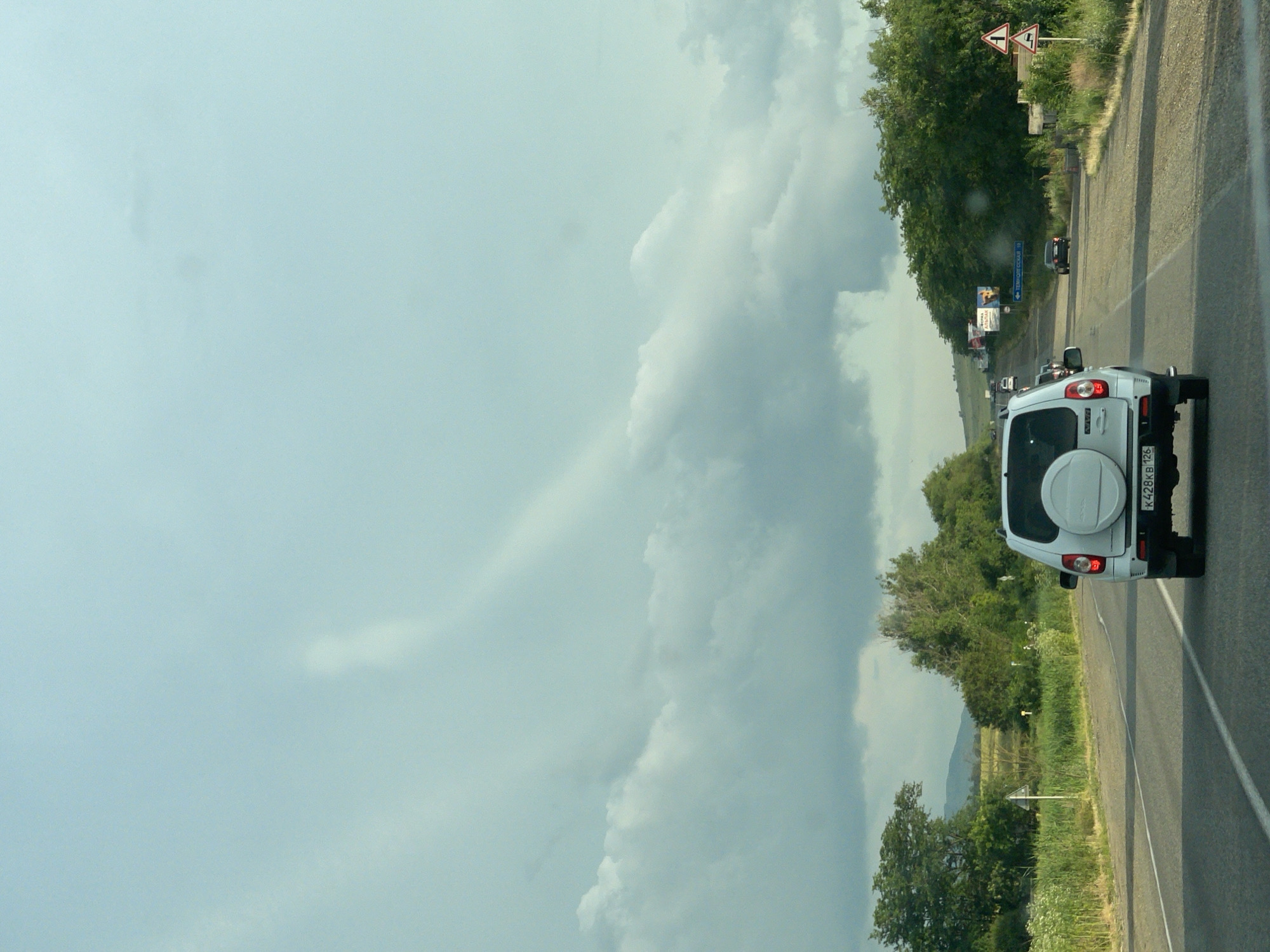 The image shows a slightly overcast day on a rural road in Tatarka, Russia. The perspective is from inside a vehicle, looking forward and slightly to the right. The primary subject in the foreground is the rear of a white SUV with a spare tire mounted on the tailgate. The license plate is visible and reads "K428KB 125". The road is asphalt and has a white line marking the lane.
To the right of the road, there are lush green trees and vegetation, with a few wildflowers visible. In the distance, more trees and a hillside can be seen. There are several traffic signs visible along the road. One is a triangular sign with two red arrows indicating a fork in the road. Another sign further down the road is blue and rectangular with text that appears to be in Cyrillic. There are other vehicles on the road ahead, including a dark-colored car and what appears to be a small bus or van. Billboards are also present along the side of the road, one of which has an image of a person.
The sky is filled with large, somewhat dramatic clouds, suggesting changeable weather. The overall impression is one of travel through a scenic, rural landscape. The time of day appears to be daytime, likely late morning or afternoon, given the light conditions. There are no people directly visible in the frame.