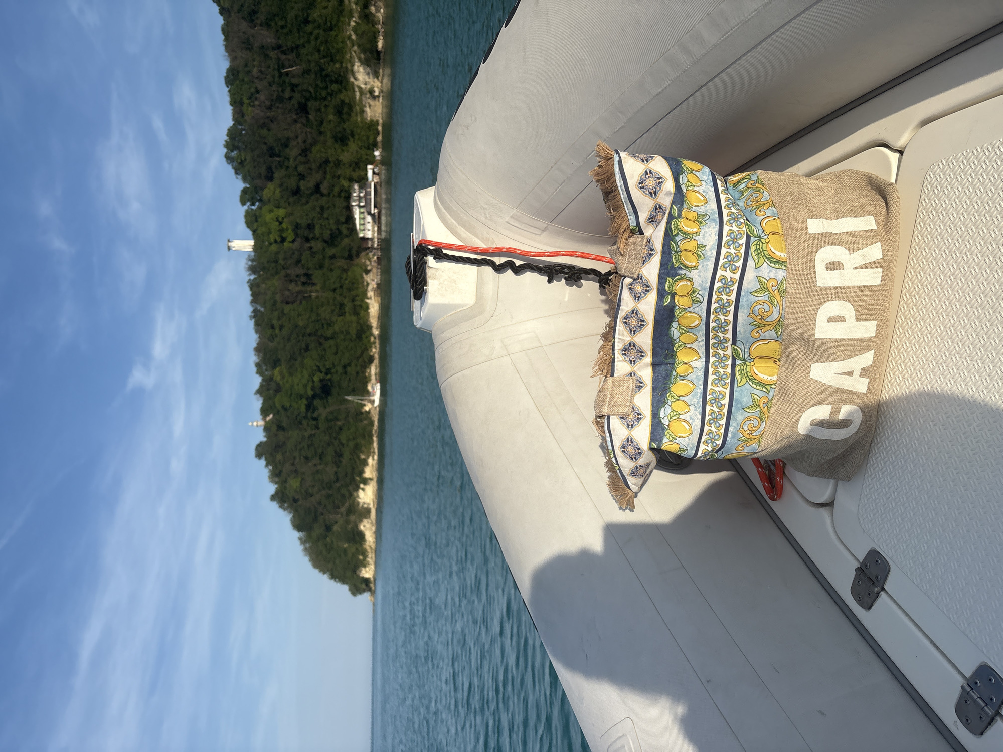 A low-angle, eye-level forensic image captures the bow of a white rigid inflatable boat (RIB) or similar small vessel, facing a distant forested coastline under a clear blue sky. In the foreground, centered on the boat's bow deck, rests a light brown burlap-style tote bag with the word "CAPRI" printed in large white capital letters. The upper section of the bag features a colorful pattern of yellow lemons, green leaves, and blue and white decorative tiles, bordered by a frayed, natural fiber trim.
A black rope and a thinner red rope, the latter appearing coiled or elastic, are secured to a white cleat or mooring post at the apex of the boat's bow. The boat's white interior surfaces, including the deck and inflatable tubes, exhibit a textured, non-slip pattern. A dark, elongated shadow, possibly cast by a person or object, extends from the left frame across the boat's port side.
The midground reveals calm, turquoise-blue water with gentle ripples. Beyond the water, a verdant, tree-covered landmass forms a distant shore, with light-colored rocky cliffs visible at its base. On the left side of the distant shore, a small white sailboat is visible on the water. Towards the center-right of the landmass, a multi-story white building is partially obscured by trees. Further right and higher on the hill, a tall, white, cylindrical structure, possibly a lighthouse or tower, stands prominently amidst the foliage, with a smaller, similar white structure visible to its right. The sky overhead is a light blue with scattered, wispy white clouds. The lighting suggests natural daylight, possibly morning or late afternoon, indicated by the distinct shadows.