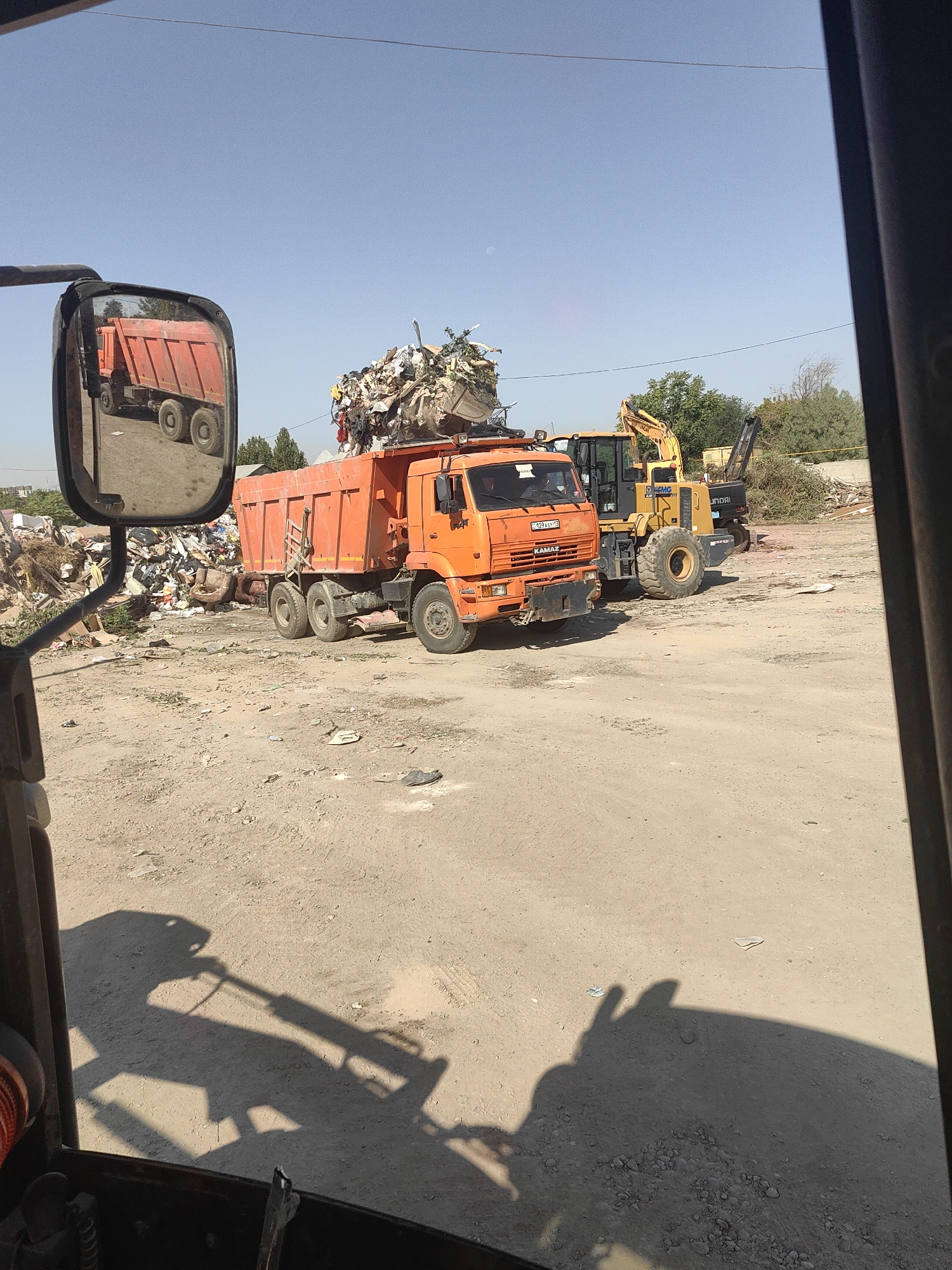 The image captures a bright, sunny day at a scrapyard in Shymkent, Kazakhstan. In the foreground, partially visible from inside a vehicle, is a side mirror reflecting an orange dump truck laden with debris. The main scene features the orange dump truck, identified as a KAMAZ with license plate 0129AEY 75, also filled with trash. Beside it, a yellow Hyundai wheel loader is parked. The ground is unpaved and sandy, littered with various types of waste, including plastic, metal, and general refuse. Trees and some buildings are visible in the background, suggesting an industrial or waste management area. The sky is clear and blue.