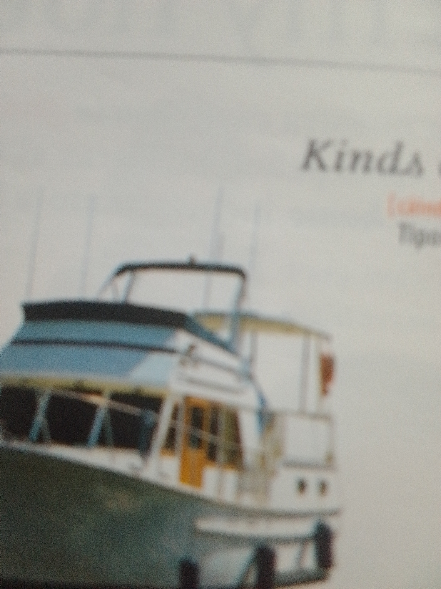 This is a blurred, low-resolution image featuring a white and teal motor yacht. The yacht has a dark blue railing and a light blue upper deck. A yellow door is visible on the side of the upper deck. Two vertical white poles extend upwards from the deck. The background is a plain, light color, likely a wall or sky. Some text is visible in the upper right portion of the image, with the word "Kinds" clearly legible, followed by partially visible words in a smaller font. The image appears to be a photograph, possibly of a brochure or magazine page. There are no people or discernible weather or time of day cues. The location context is La Tagua, Venezuela.