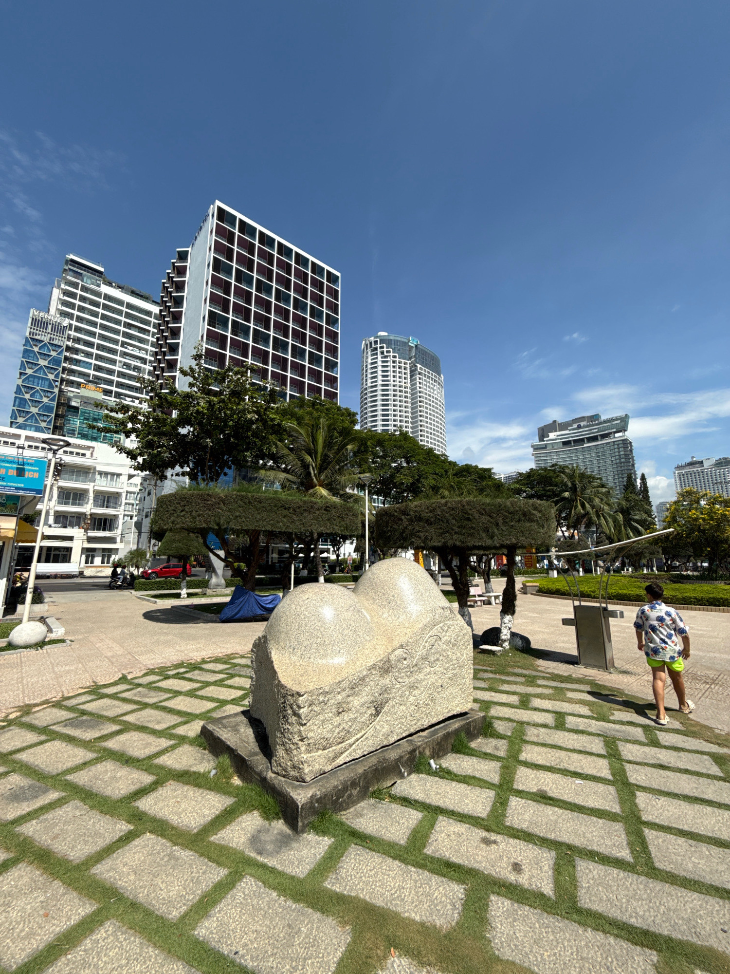 The image depicts an outdoor scene in the city of Nha Trang, Vietnam, likely near the coast given the presence of modern high-rise buildings and palm trees. In the foreground, a large, light-colored stone sculpture, resembling a cluster of smooth rocks, is situated on a raised concrete base. The sculpture is surrounded by a paved area with a grid pattern of square stones interspersed with patches of grass. To the right of the sculpture, a young person wearing a colorful patterned shirt and lime green shorts walks away from the camera.
In the mid-ground, several trees are visible, some with elaborately sculpted canopies, adding to the urban park aesthetic. A blue tarp is visible partially covering something on the ground behind the sculpture. In the background, a street runs horizontally, with a few vehicles, including a red car and motorcycles, and a pedestrian walking on the sidewalk. Further in the background, a collection of modern, multi-story buildings dominates the skyline. These buildings vary in architectural style and height, with one particularly tall, white building featuring a grid pattern of dark windows and balconies. The sky above is clear and blue with a few wispy clouds, indicating a sunny day. The overall scene suggests a public space within a developed urban environment, likely a park or promenade area in Nha Trang.