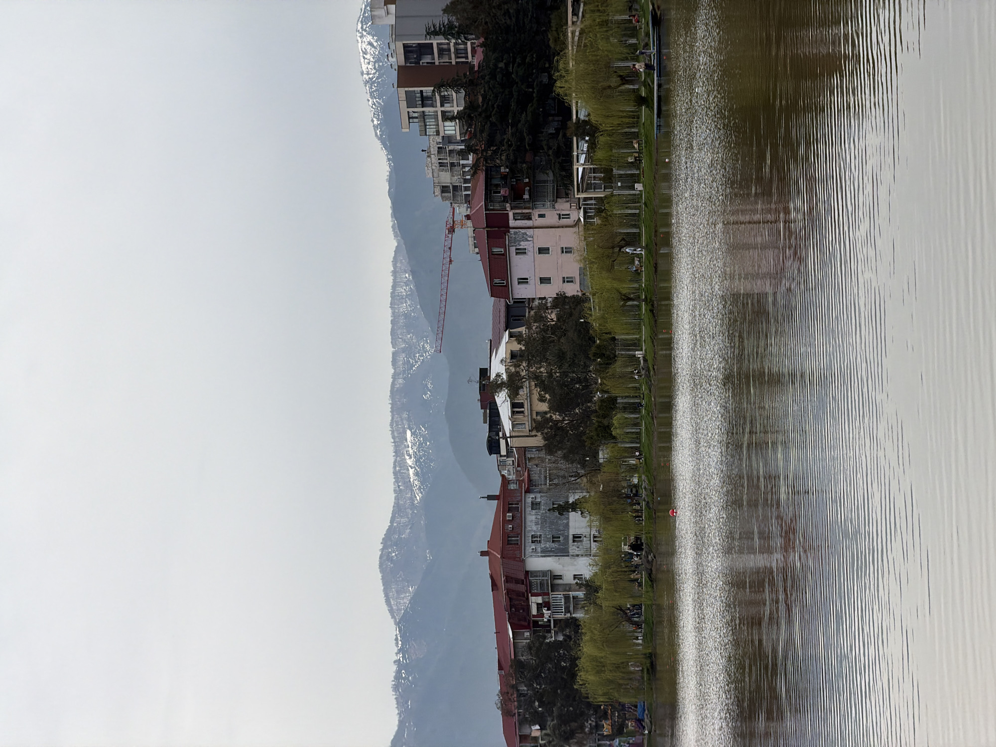 The image shows a body of water reflecting buildings and mountains. On the right, the water's surface is rippled. Along the shoreline, there are trees. Buildings of various heights and architectural styles are adjacent to the water. A construction crane is visible among the buildings. In the background, mountains with snow-capped peaks rise into a light gray sky. It is the reflection of the lake in Batumi, Georgia.