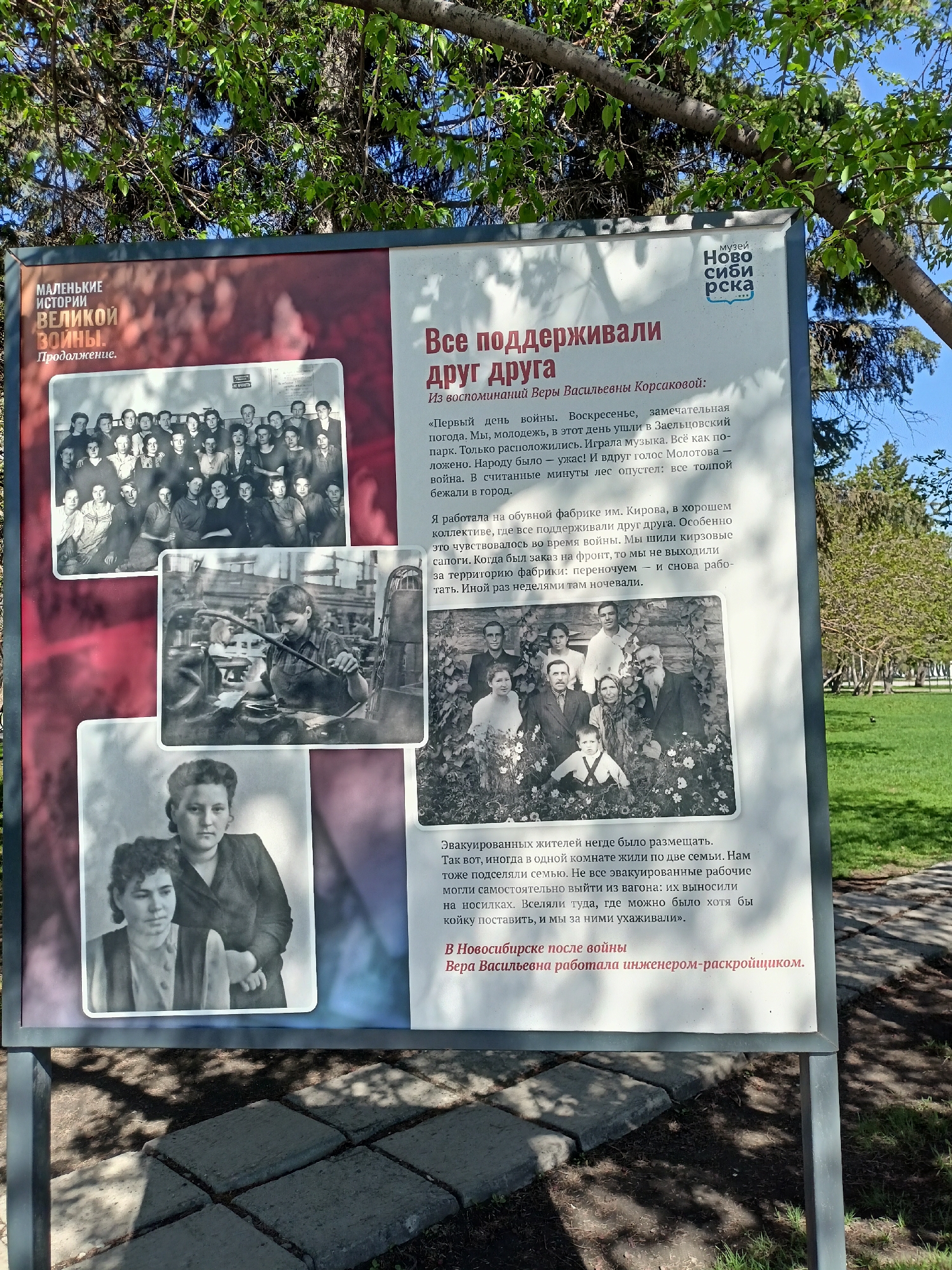The image shows an outdoor information sign in Novosibirsk, Russia, located in a park or other green space. The sign features the title "Все поддерживали друг друга" (Everyone Supported Each Other) and is part of a series titled "Маленькие истории Великой Войны" (Small Stories of the Great War). It displays excerpts from the memories of Vera Vasilyevna Korsakova, including recollections of the first day of the war and work at the Kirov shoe factory. The sign also includes black-and-white photographs depicting people and scenes related to these memories, such as a group of young people, a worker in a factory, and a family portrait. The background includes trees, grass, and a stone path. The logo of the "Музей Новосибирска" (Museum of Novosibirsk) is visible on the sign.