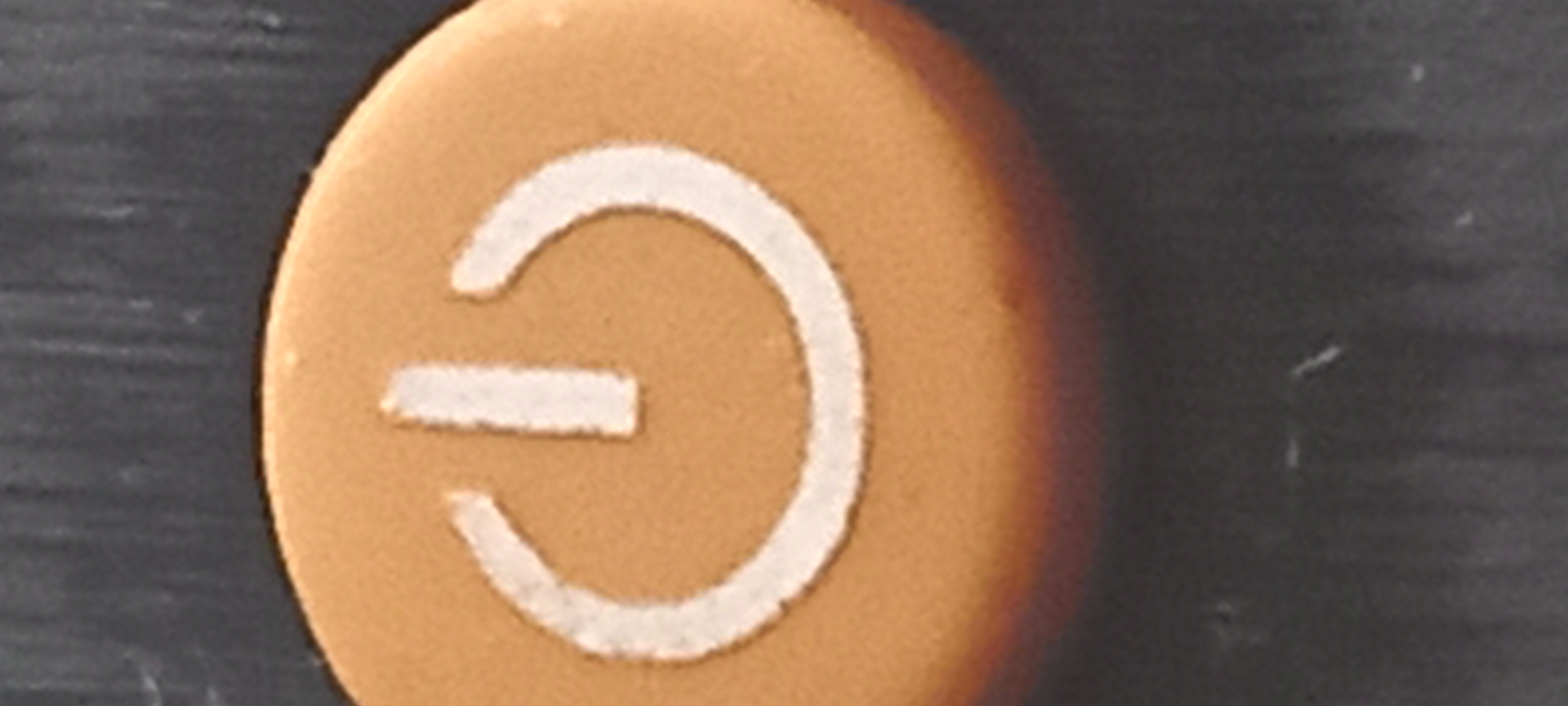 The image shows a close-up of a round, tan-colored button with a white power symbol on it. The symbol consists of a white circle, broken at the top, with a horizontal white line inside the circle. The button is sitting on a dark, textured surface. The background is out of focus. There is no information to identify this object's location as Kryvyi Rih, Ukraine.