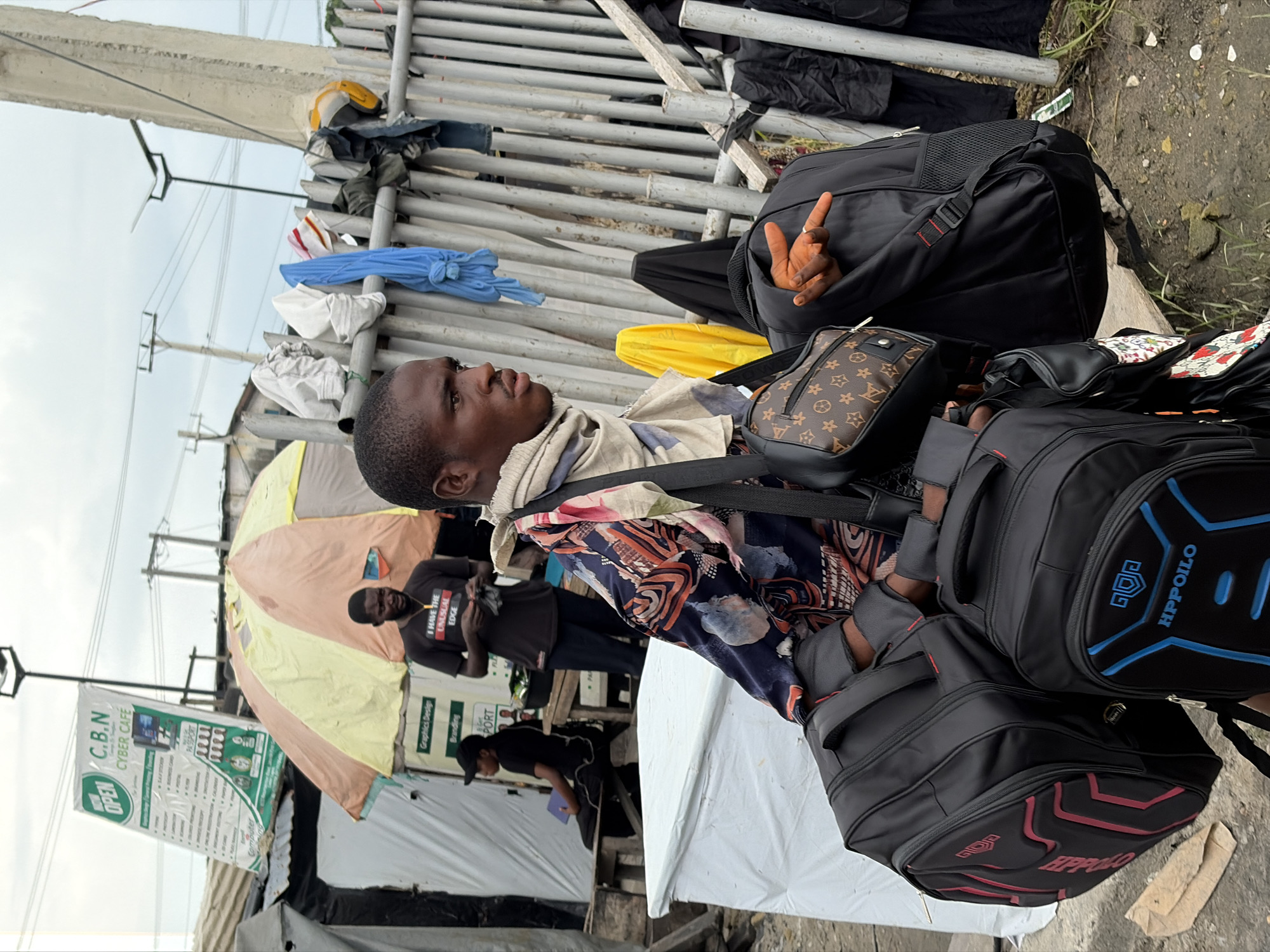 A young Black male is depicted from the waist up, facing right, in an outdoor setting in Igboefon, Nigeria. He is carrying several black backpacks and a small brown Louis Vuitton-patterned bag. He wears a light-colored scarf around his neck and a dark patterned shirt. His right hand is raised, with fingers spread. In the background, a sign for "C.B.N CYBER CAFE" is visible, along with a yellow and orange umbrella, other individuals, and laundry hanging on a metal fence. The ground is dirt and debris.