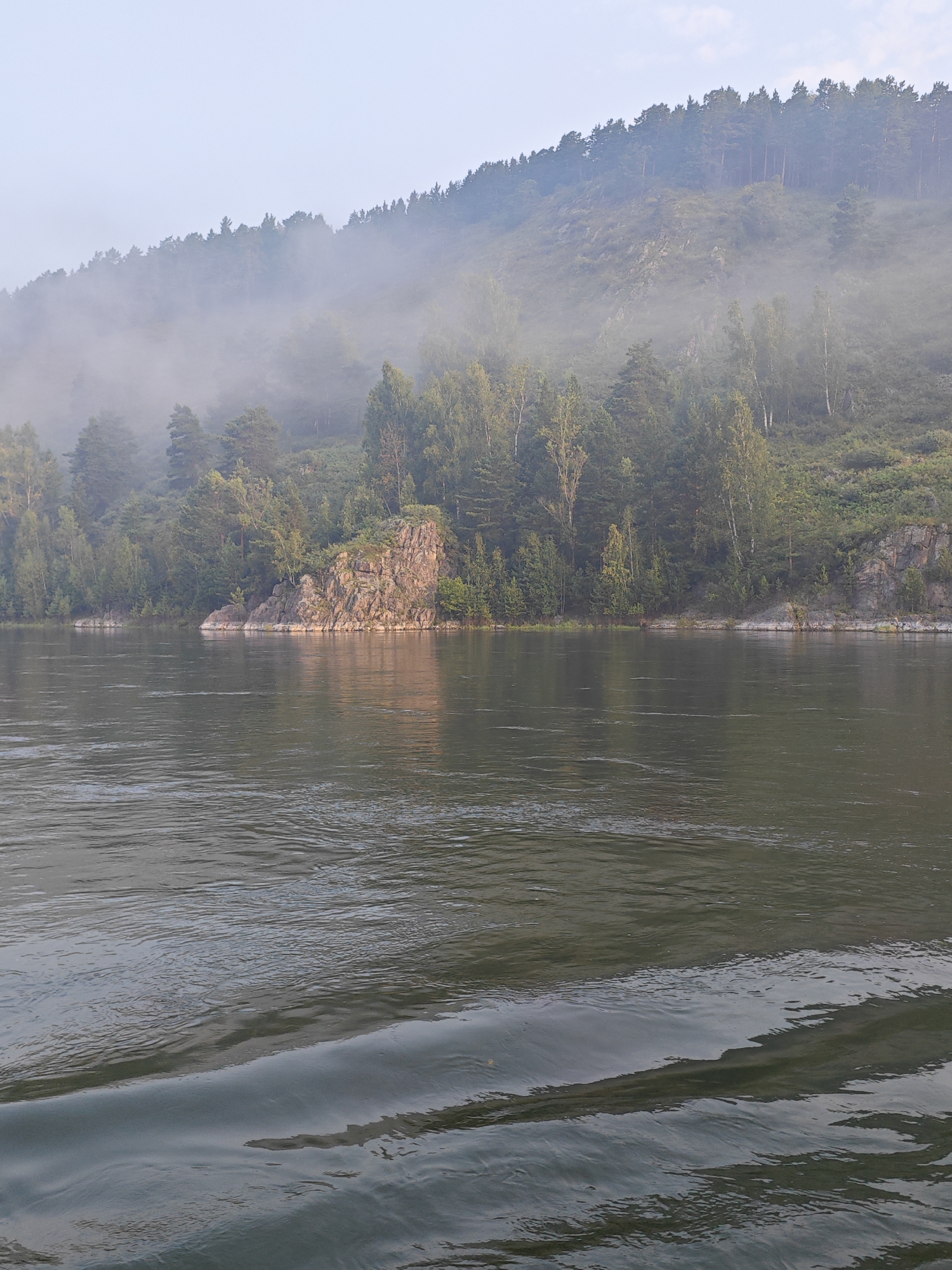 The image shows a wide, calm river flowing in front of a forested hillside. A light mist hangs over the trees, particularly on the higher slopes, giving the scene a soft, ethereal quality. The water in the foreground shows ripples, suggesting movement, perhaps from a boat or the natural flow of the river. The bank of the river is lined with trees, with a prominent rocky outcrop on the right side of the image. The sky is overcast and a pale blue. The location is the Yenisei River in the village of Atamanovo, Krasnoyarsk Krai, Russia.