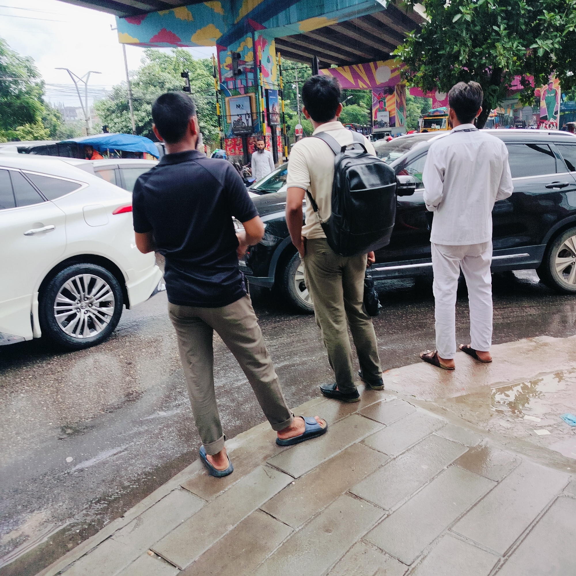 This is a street-level, eye-level, medium shot photograph taken outdoors in Dhaka, Bangladesh, under an overpass adorned with colorful murals. The scene depicts three men standing on a wet, paved surface next to a road with traffic.
On the left, a man is seen from behind, wearing a dark blue t-shirt and khaki pants, standing on a blue sandal. He is partially facing a white car. To his right, another man, also viewed from behind, wears a light beige t-shirt and khaki pants, carrying a black backpack. He is standing on dark sandals. Further to the right, a third man, dressed in a white shirt and white pants, is seen from behind, also wearing dark sandals. He is standing next to a black SUV.
The road surface is wet, indicating recent rain, with visible puddles and reflections. The overpass structure above features bright, abstract murals. Trees and foliage are visible in the background, along with traffic. The overall atmosphere suggests a bustling urban environment, possibly during or after a rain shower.