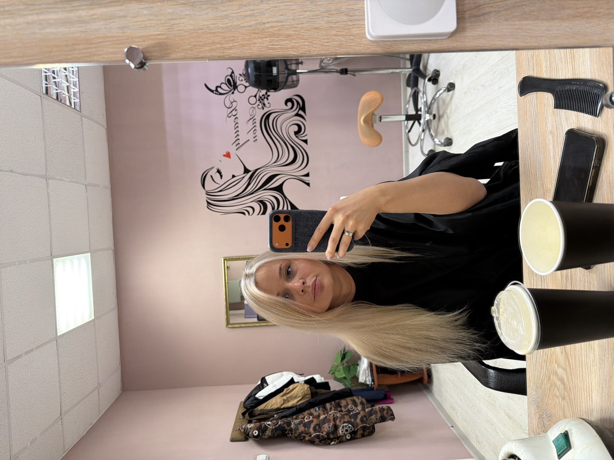 A photograph, taken in what appears to be a beauty salon in Sarapul, Russia. A blonde woman is taking a selfie in a mirror. The mirror reflects the interior of the salon, including a wall decal of a woman's face and flowing hair, a salon chair, and a wall. The table near the mirror holds a comb, a phone, two black cups with white lids, and a white object with a green button. In the room, a pile of clothing and a plant are visible on the floor. The ceiling of the room has a grid of square lights. The walls are painted in a light pink color.