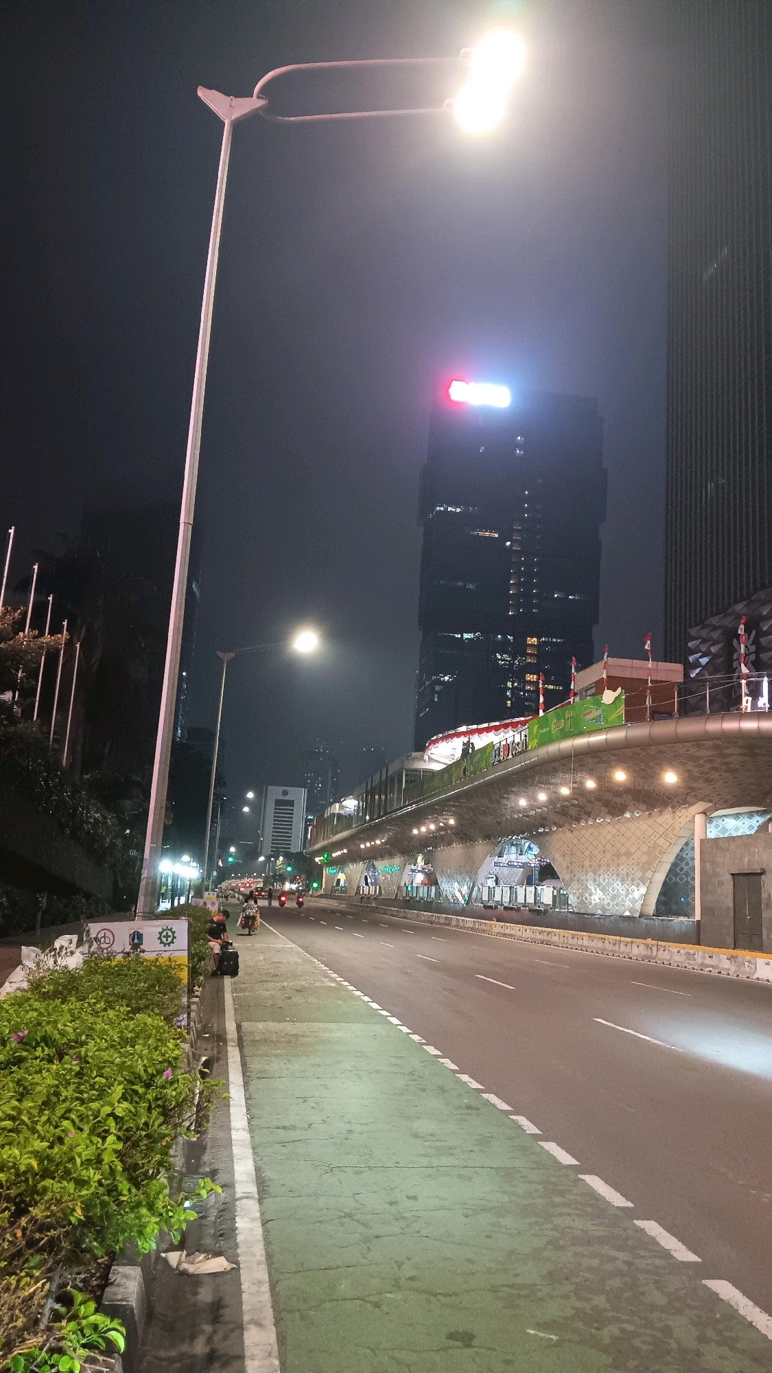 The image depicts a nighttime street scene in Central Jakarta, Indonesia, likely near Gelora Bung Karno (GBK) Stadium, given the architectural style and prominence of a large building with glowing signage. A tall, modern streetlamp with two bright lights illuminates the foreground. To the left, lush green bushes line a sidewalk that transitions into a marked green bike lane. Beyond the bike lane, a multi-lane road stretches into the distance, with several motorcycles visible, some of which appear to be stationary.
In the mid-ground, a striking elevated walkway or bridge features intricate decorative tiling on its underside and is lined with lights. Several flags, including Indonesian flags, are visible on this structure. Behind it, a very tall, dark skyscraper dominates the skyline, its upper floors adorned with bright red and white illuminated signage that appears to spell out a word. The overall atmosphere is urban and bustling, despite the late hour, with the artificial lights cutting through the night sky. The presence of construction elements on the elevated walkway suggests ongoing development or renovation in the area.